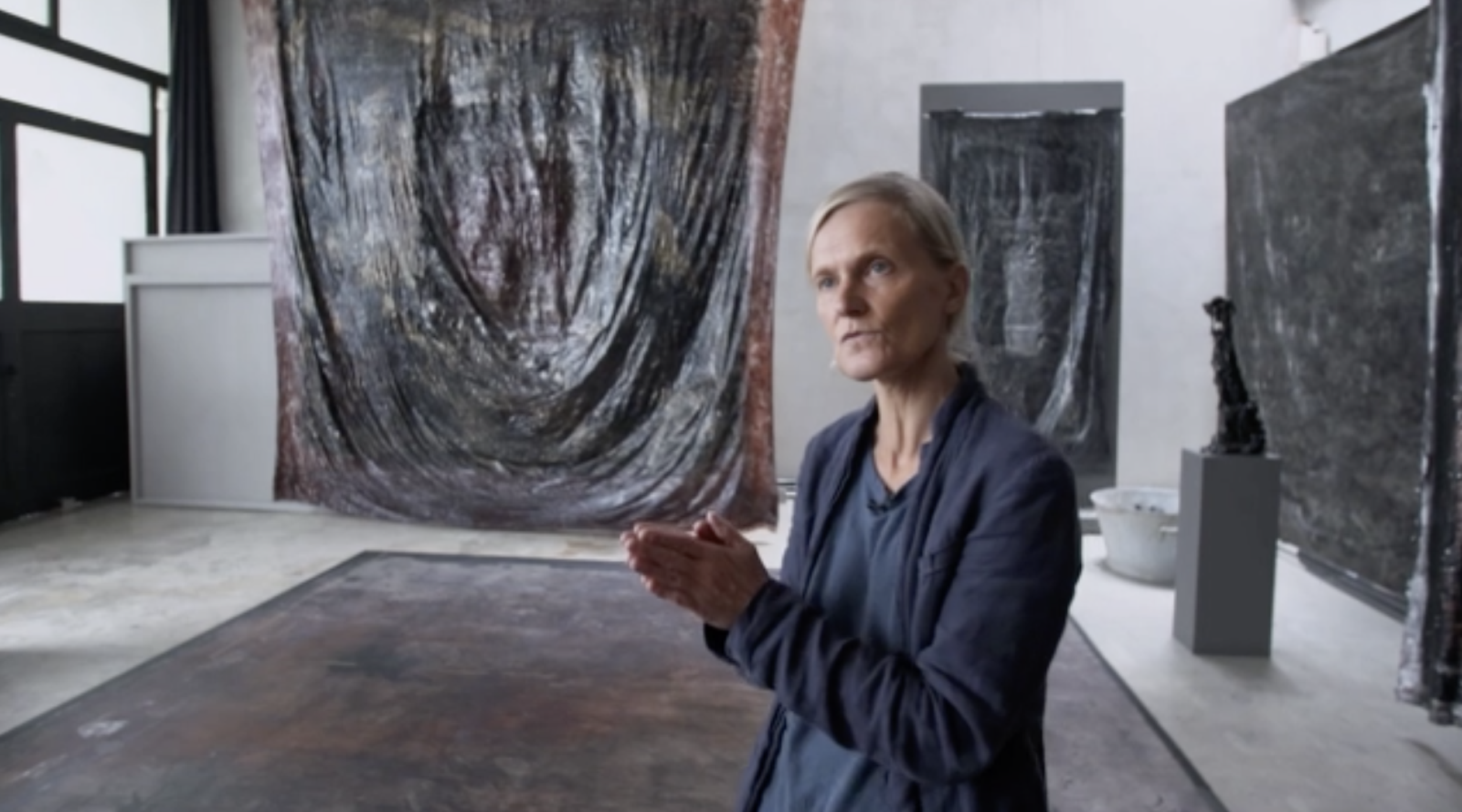 Eine ältere Frau sitzt in einem Kunststudio vor mehreren Gemälden und Skulpturen, während sie mit gefalteten Händen spricht.