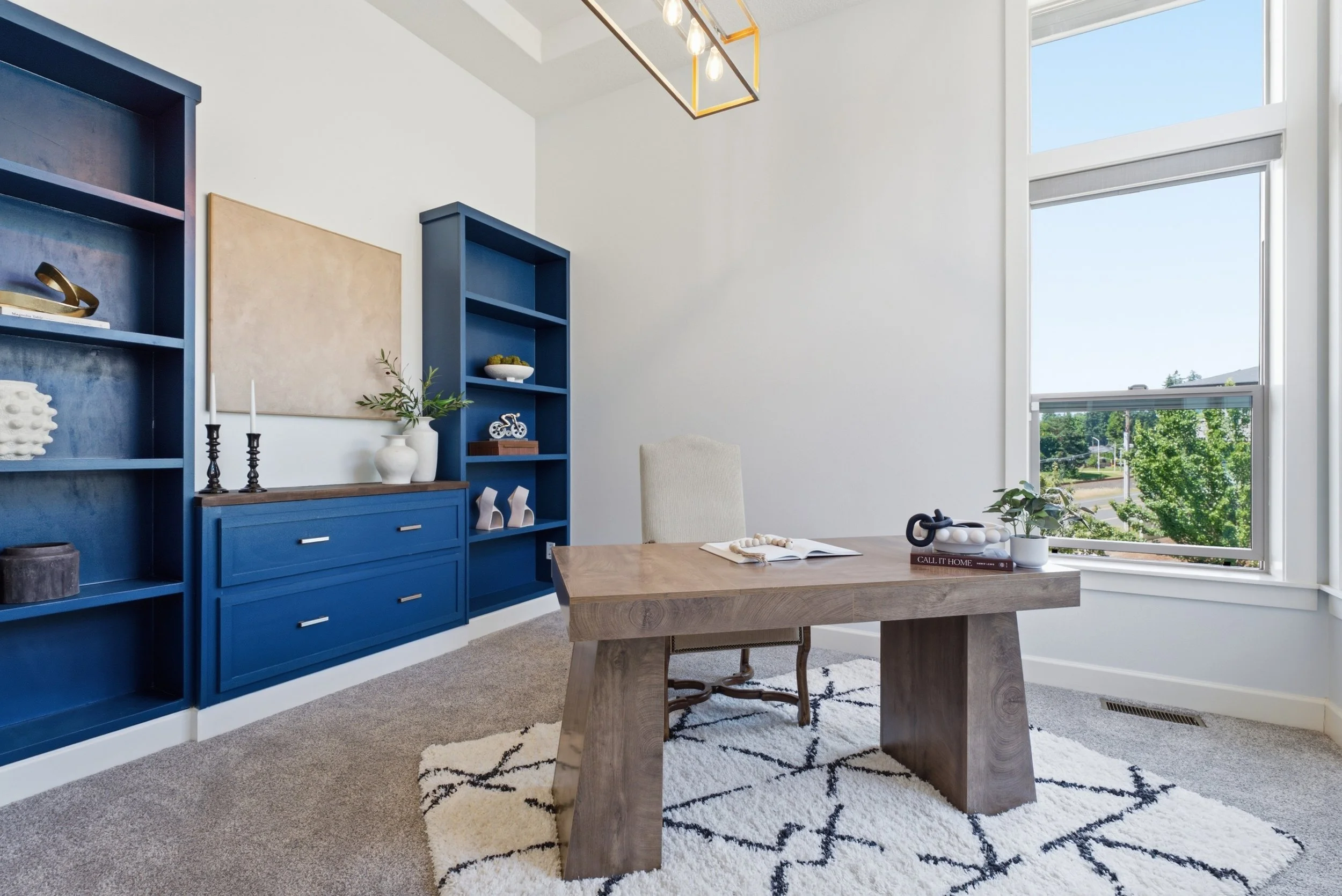 Modern dining room with a wooden table, beige upholstered chair, blue bookshelves and cabinets, decorative objects, large window with a view of trees, and a black and white patterned rug.