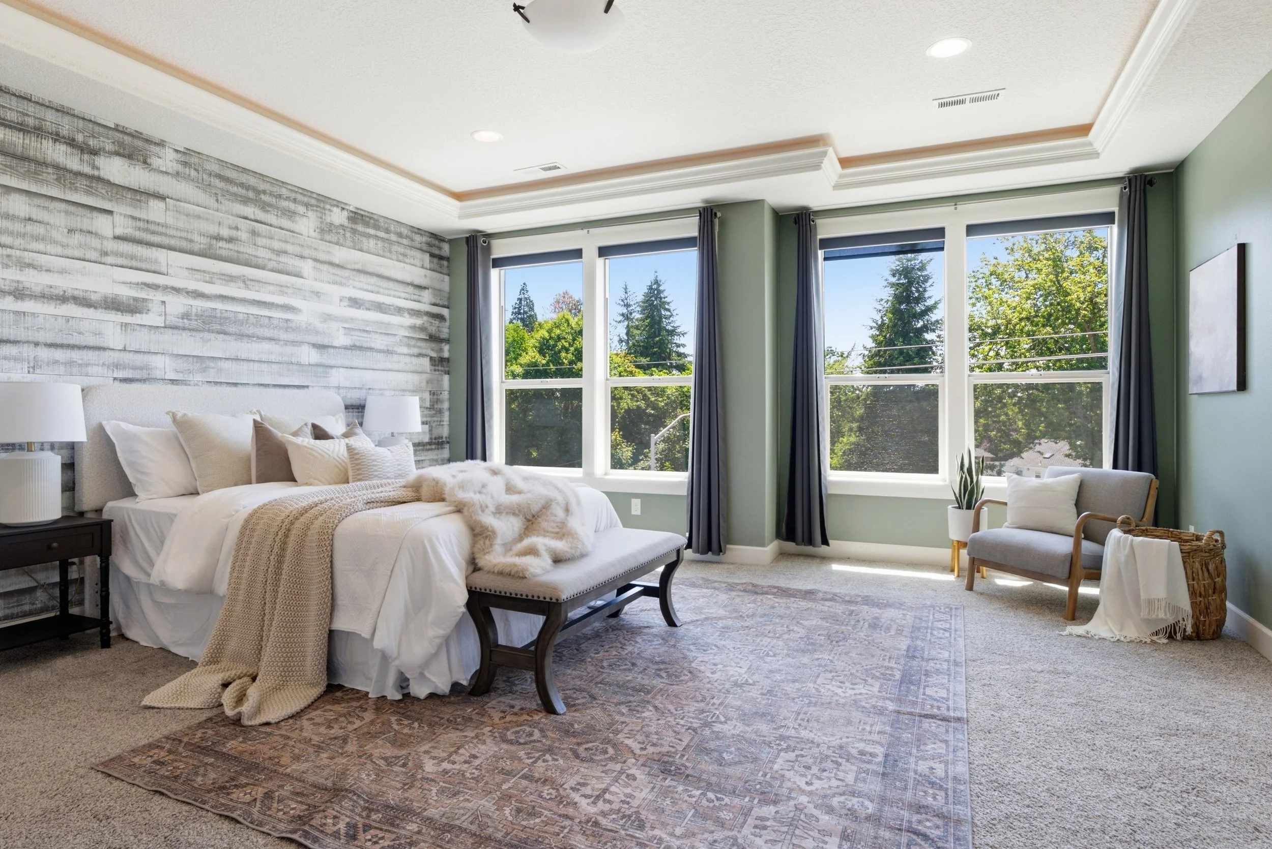 A spacious bedroom with a large bed, nightstands, lamps, a bench, and a chair, featuring a textured accent wall and large windows with a view of trees outside.