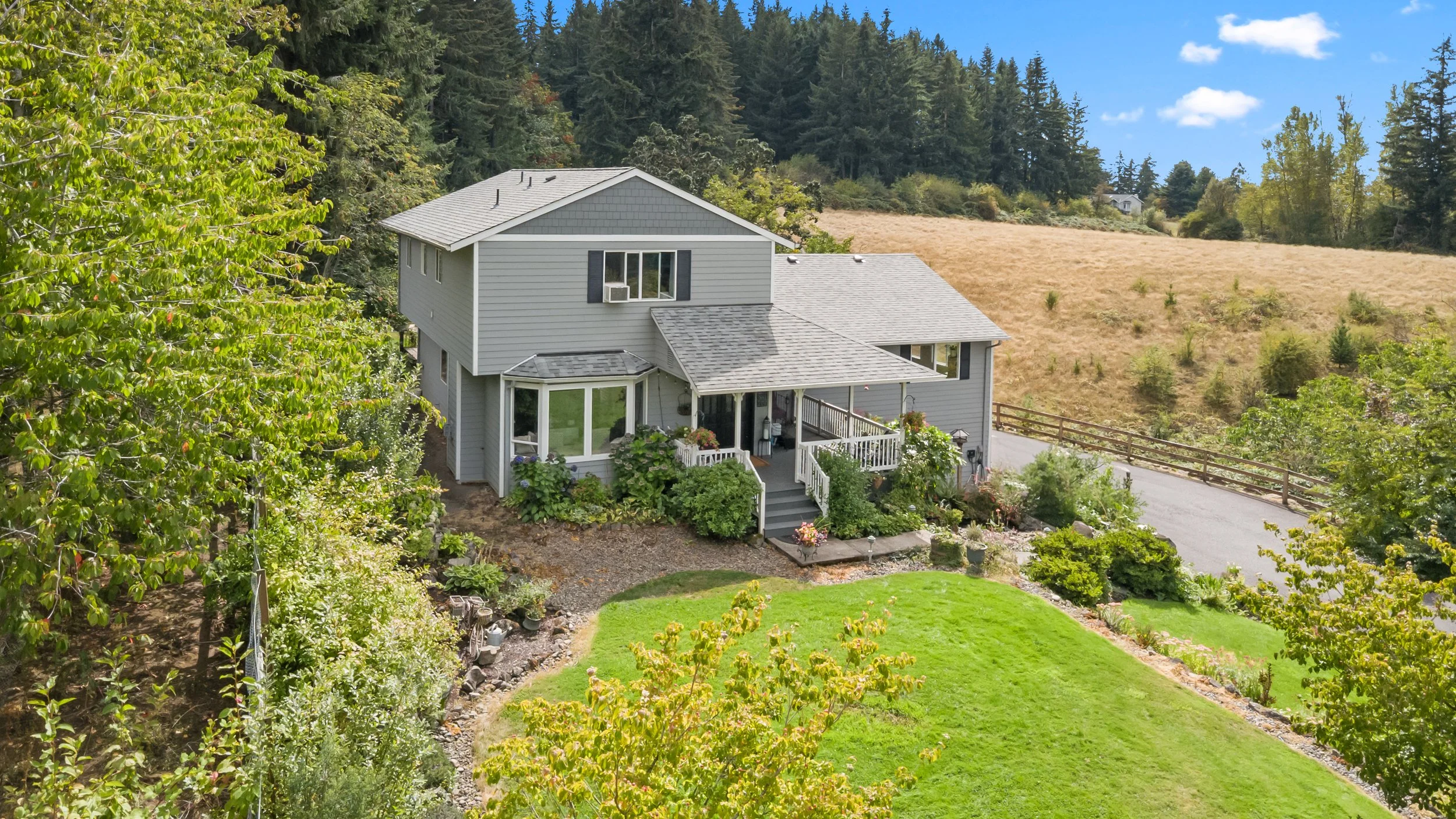 A two-story gray house with a porch and garden, situated in a lush green yard with trees, bushes, and a wheelchair ramp, set against a backdrop of a hill with dry grass and a forested area, under a blue sky with scattered clouds.