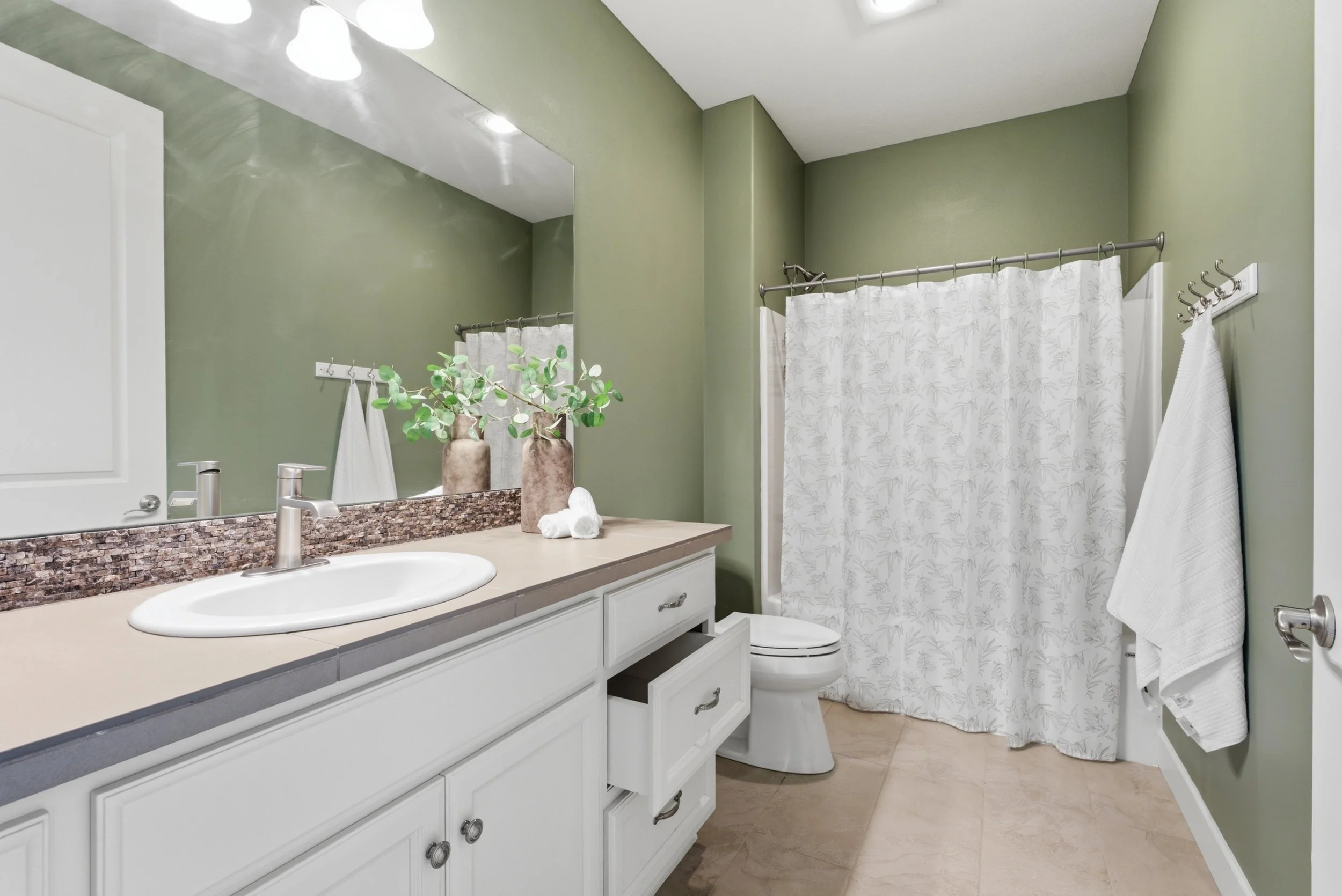 A bathroom with a green wall, white vanity with a beige countertop, a sink, and silver faucet, decorative vases with greenery, a mirror with four lights, a white toilet, a shower with a patterned curtain, white towels, and a towel rack.