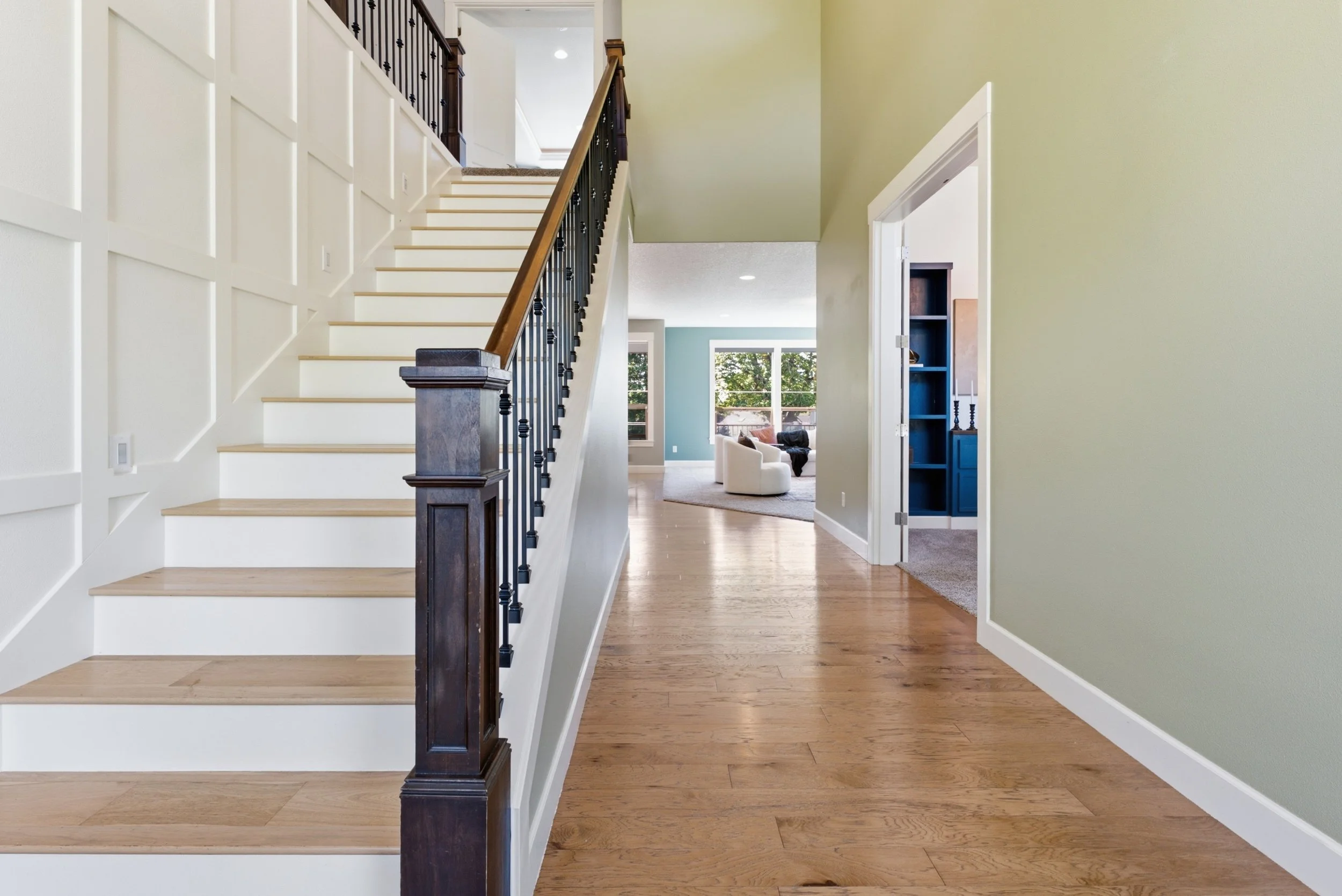 Interior view of a home with a staircase on the left, an open doorway on the right leading to a room with blue walls and built-in shelves, and a living room visible in the background with large windows, a white armchair, and a black armchair.