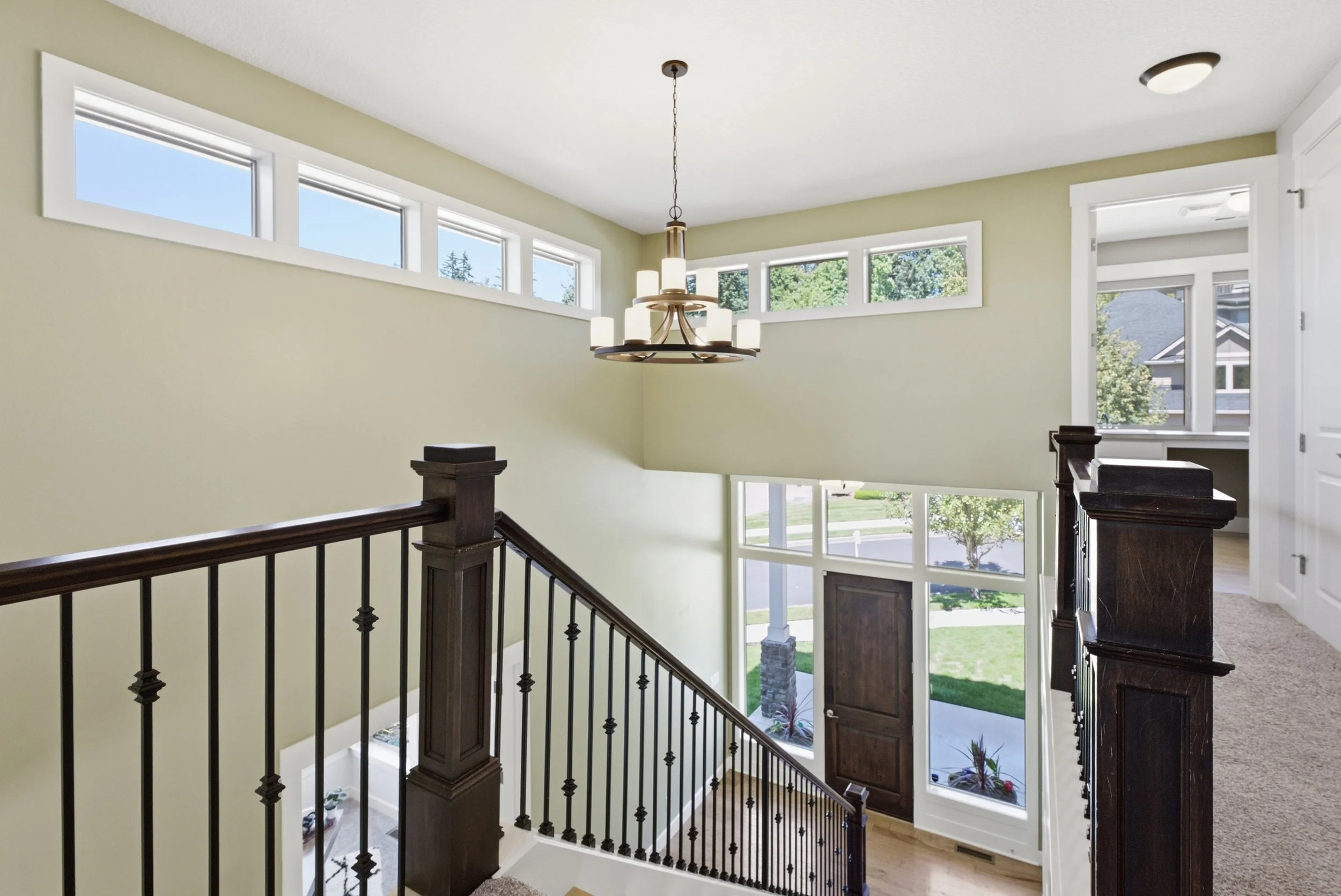 View of a bright interior staircase landing with large windows, a chandelier, and a door leading outside to a yard with trees.