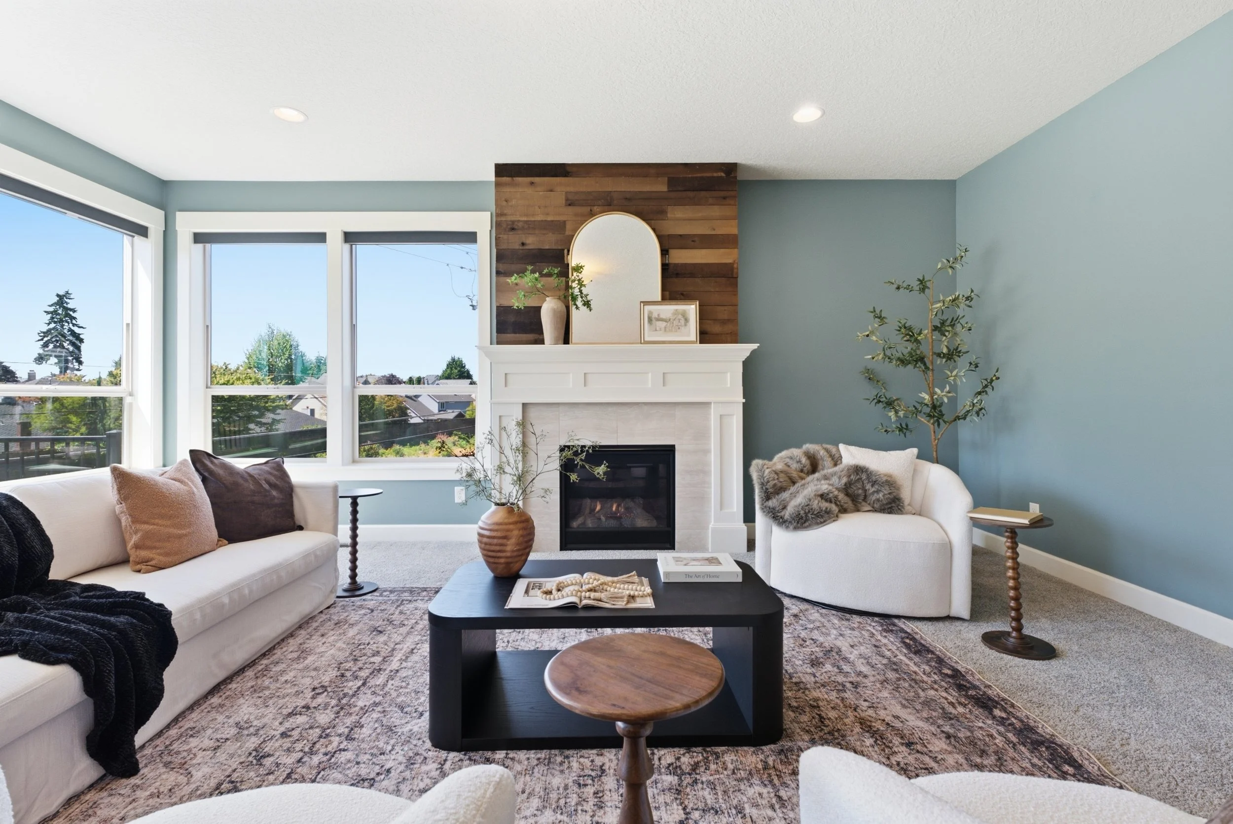 Living room with a white fireplace, large windows, and a mix of modern and rustic decor including sofa with pillows, a black coffee table with books and a small wooden stool, a patterned rug, and plants.