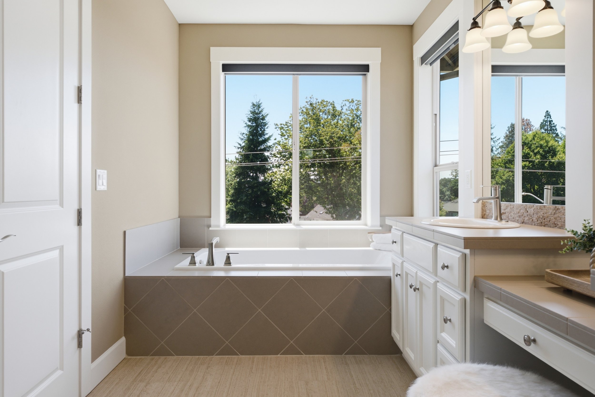 Bathroom with a large window showing trees outside, a bathtub, white cabinetry, and a sink, with beige and brown tones.