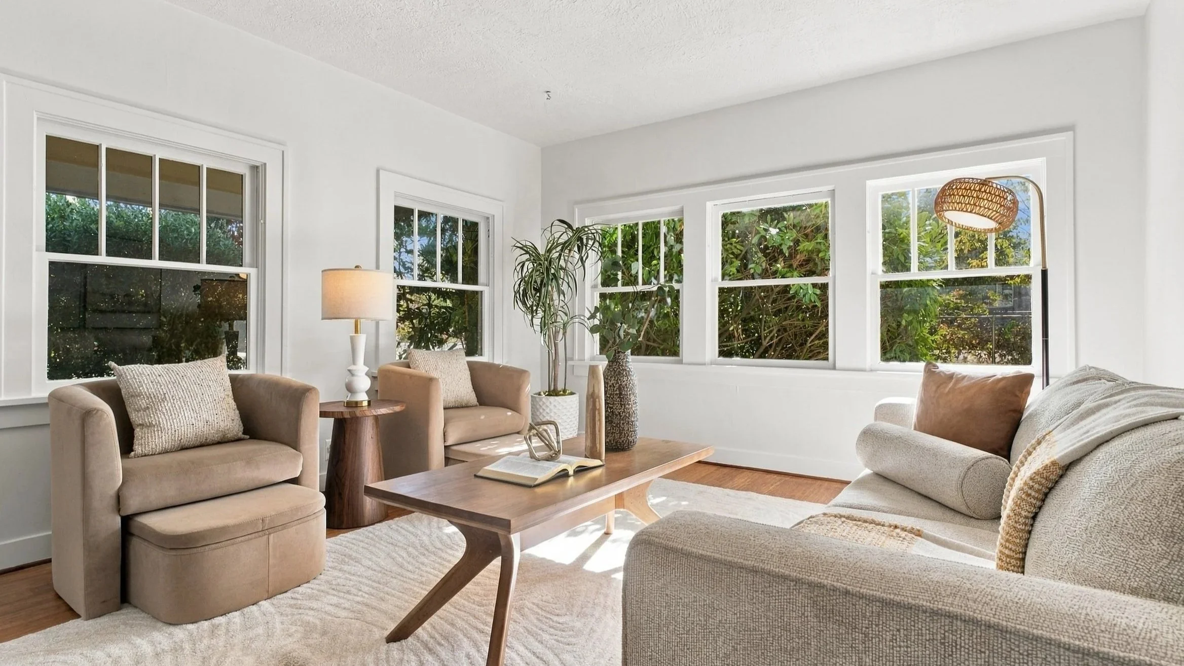 Living room with beige sofas, a wooden coffee table, large windows, and indoor plants, illuminated by natural light.