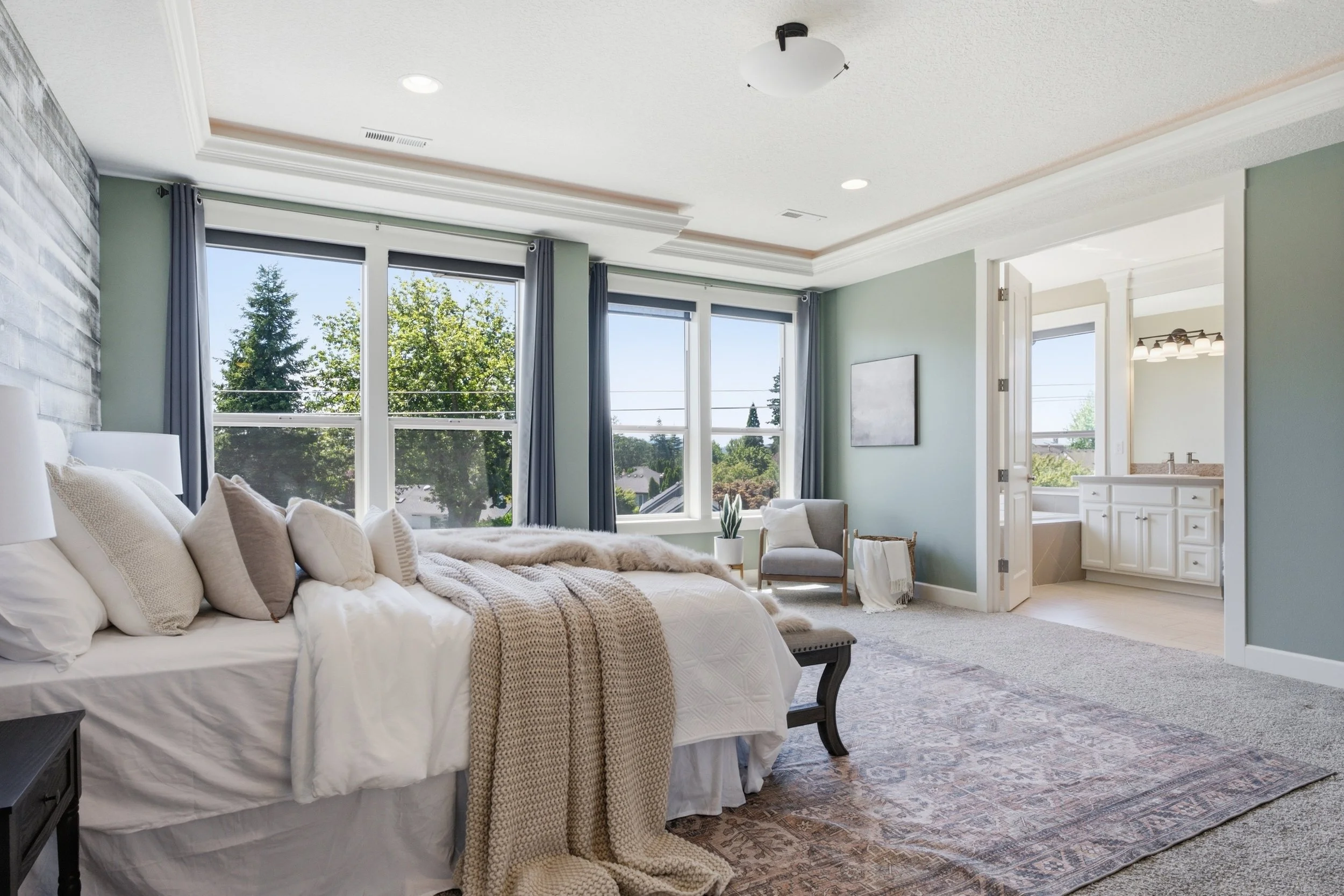 A bedroom with a large bed covered in white bedding and beige pillows, a textured beige throw blanket, a small potted plant on a chair, and three large windows with dark curtains showing trees outside. The room has a light green wall, crown molding, 