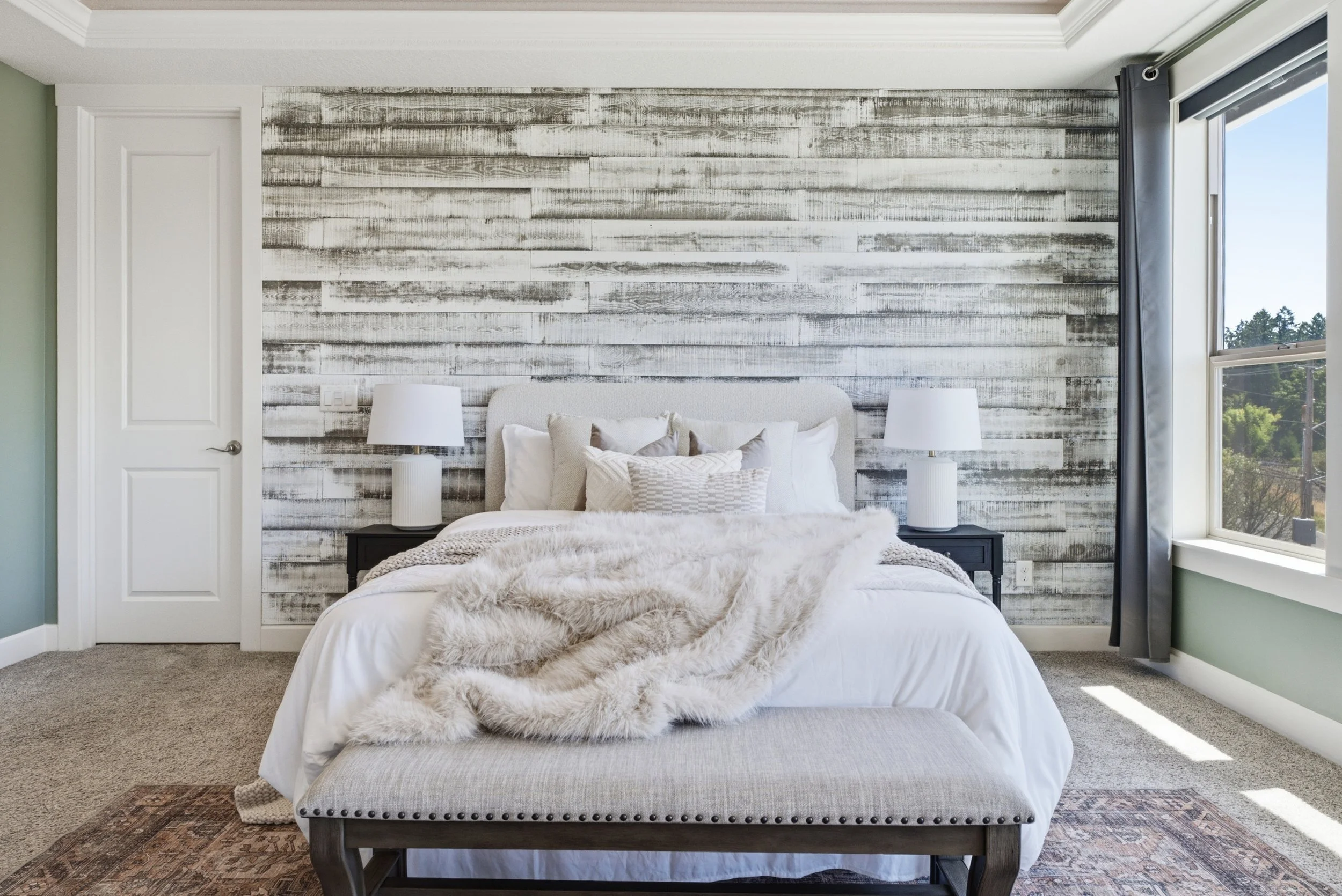 A bedroom with a bed, white bedding, and a faux fur throw. The wall behind the bed has a weathered wood plank design. There are two white lamps on black nightstands, and a window with gray curtains on the right.