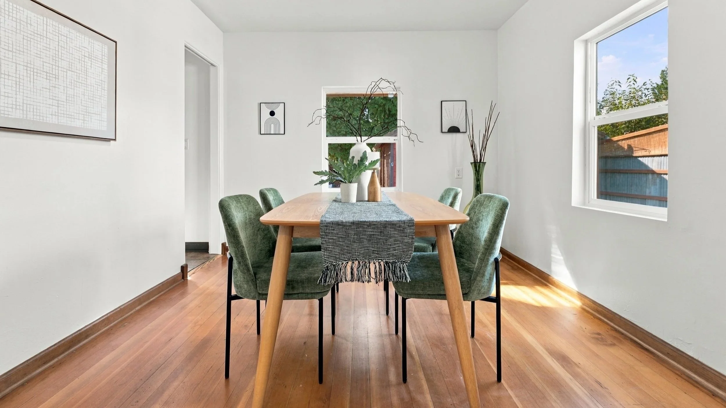 A dining room with a wooden table, six upholstered green chairs, a table runner, and decorative vases with branches and greenery. White walls with framed artwork, hardwood floors, and a large window with a view of a fence and trees.