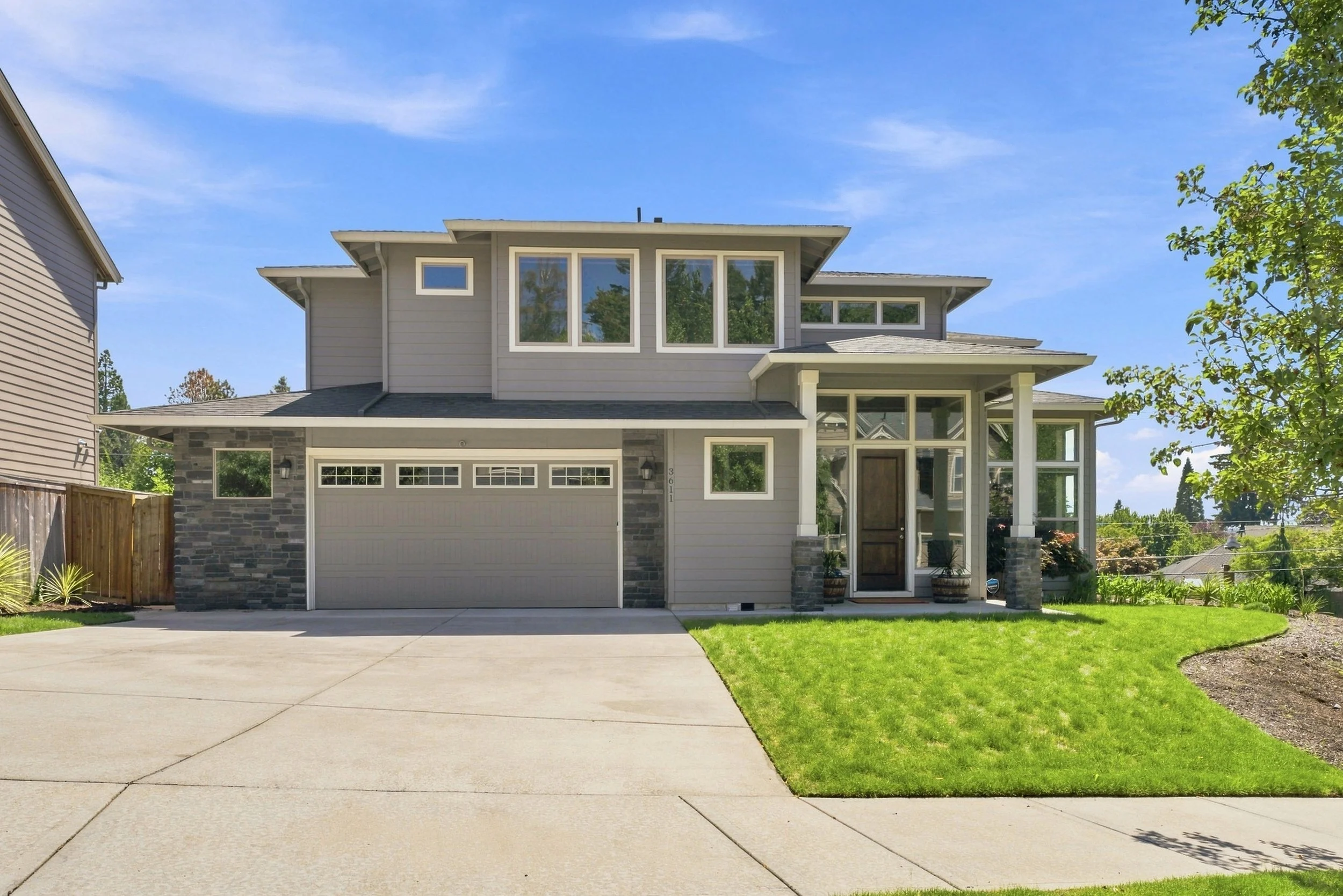 A modern two-story house with grey siding, large windows, a two-car garage, a small covered porch with potted plants, a well-manicured lawn, and a blue sky with scattered clouds.