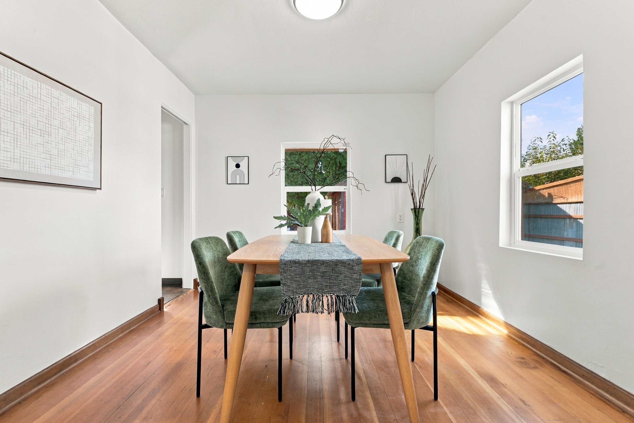 A dining room with a wooden table surrounded by six green upholstered chairs, a large white vase with leafy branches, two framed black and white artworks on the wall, and windows letting in natural light.