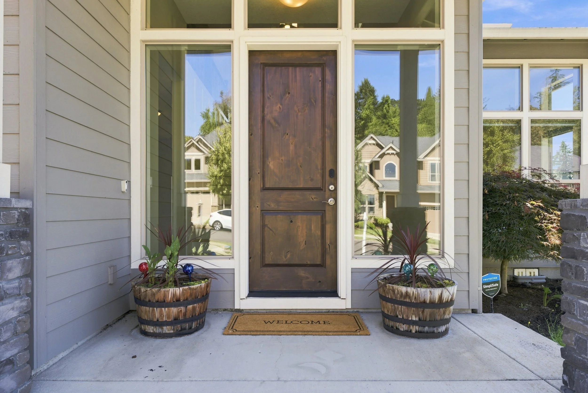 Front porch of a house with a wooden door, two large potted plants with colorful garden ornaments, a brown welcome mat, and a security system sign. The house has beige siding and large windows.