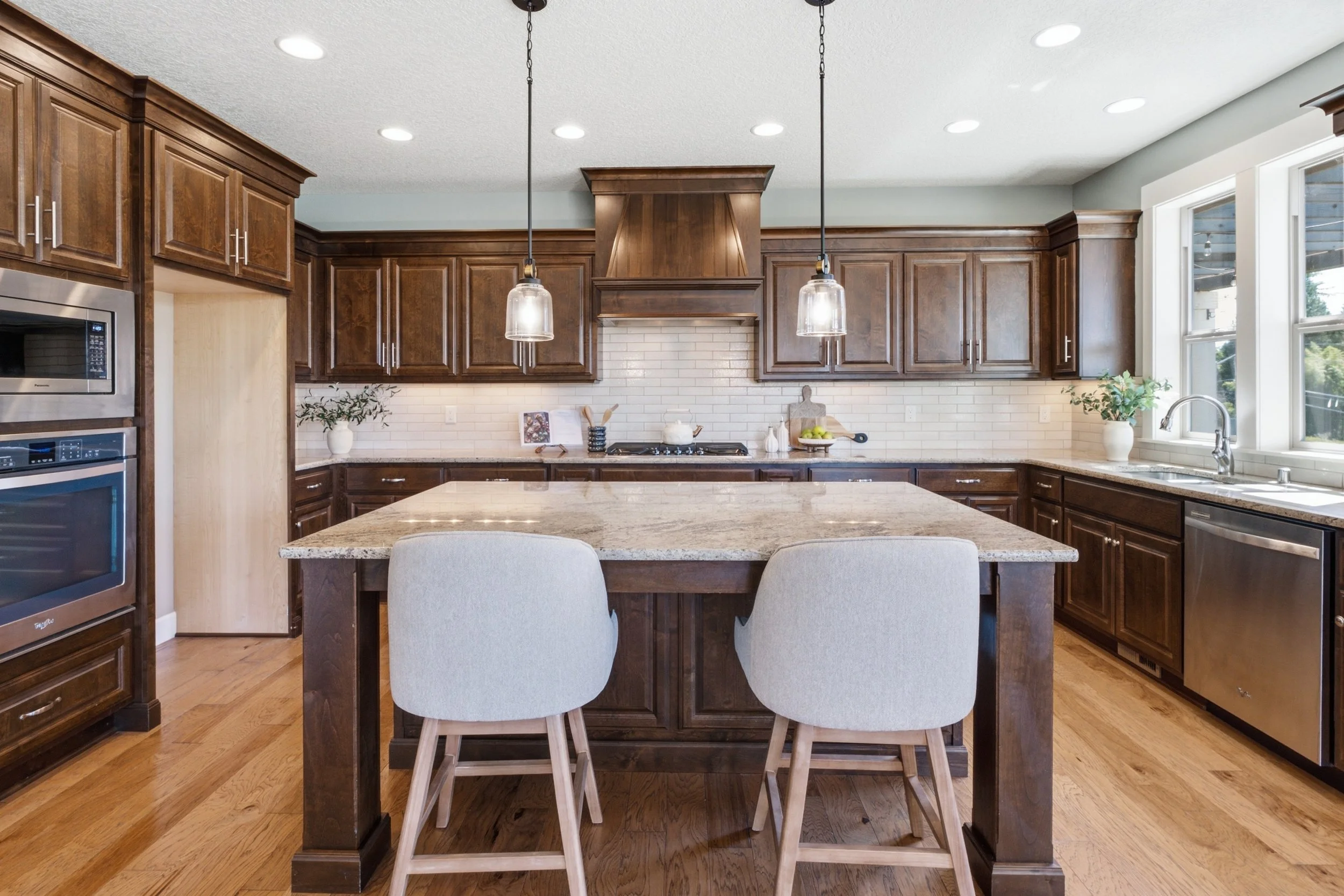 A modern kitchen with wooden cabinets, a large island with a granite countertop, pendant lights, and a window with a view outside.