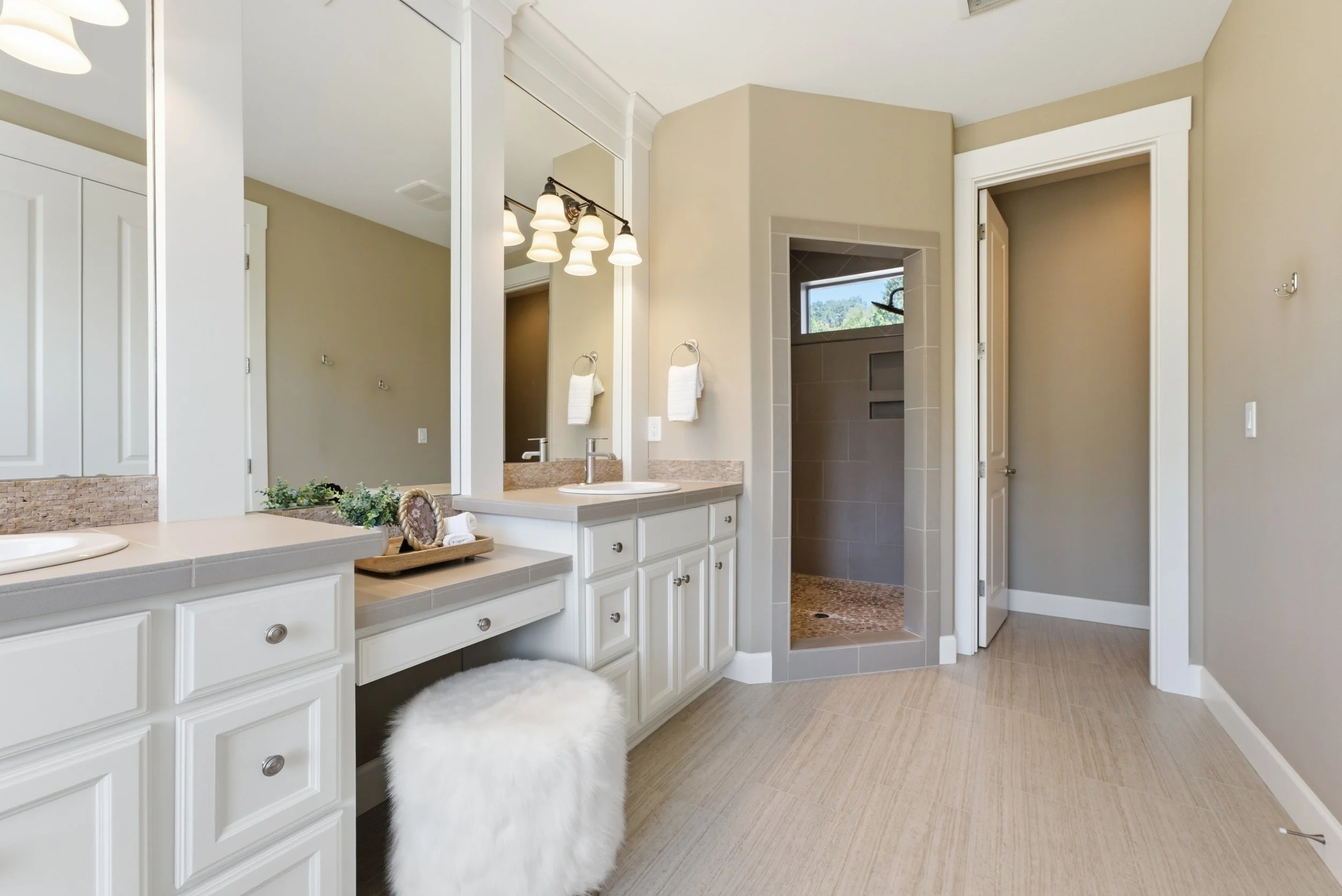 Bright bathroom with large mirror, double vanity, beige walls, white cabinetry, and a shower with gray tiles, and a small window showing greenery outside.