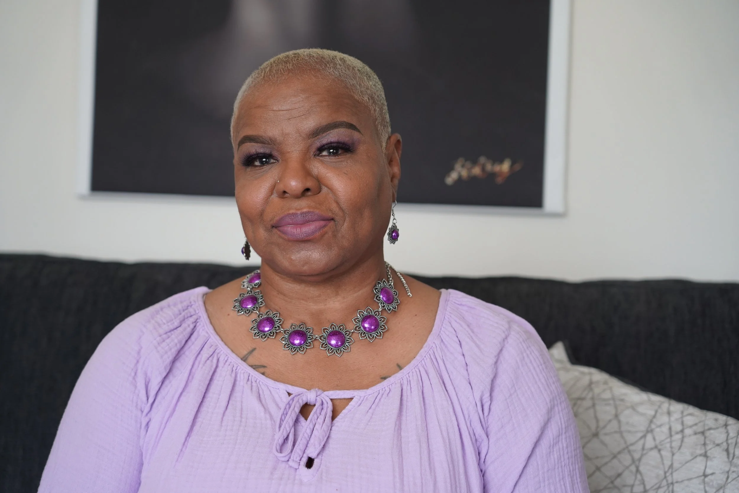 A woman with short blonde hair wearing purple eyeshadow, purple jewelry, a purple top, sitting on a black couch, with a black framed picture on a white wall behind her.