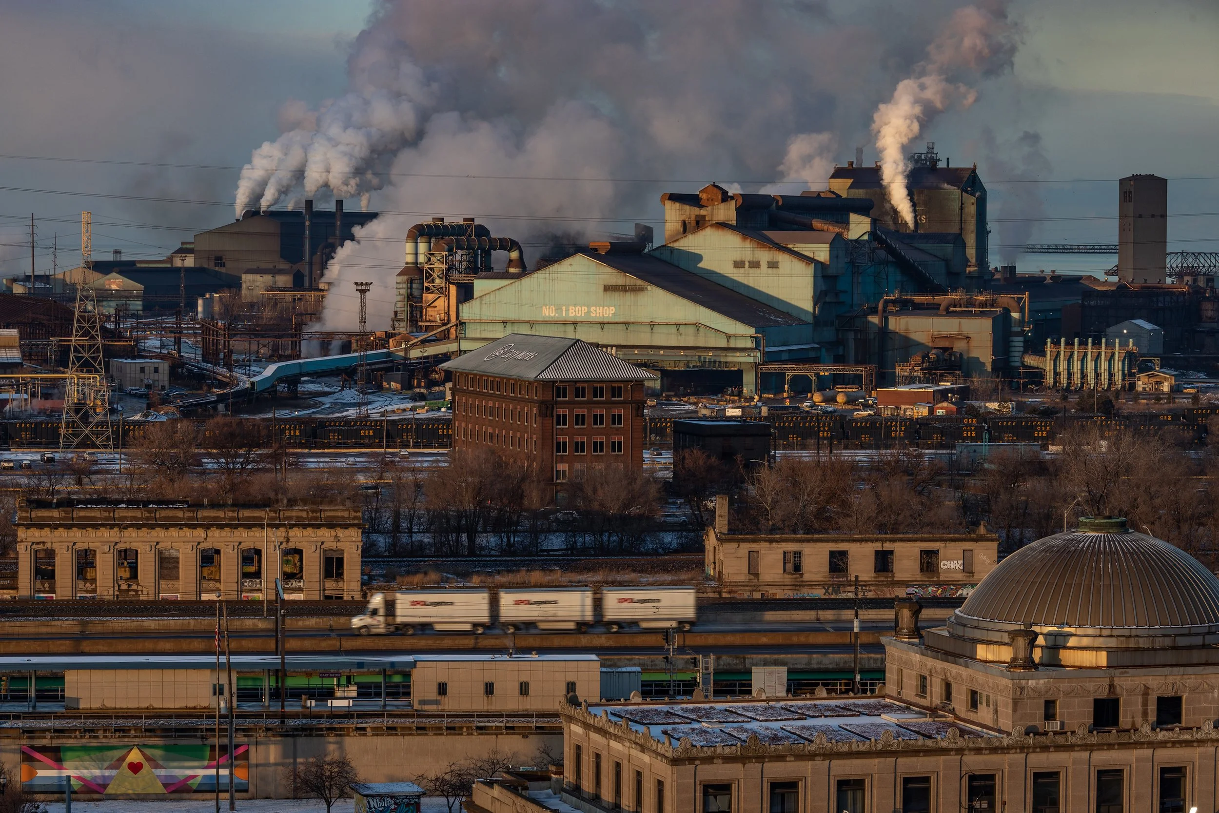 Industrial factory with smoke rising from chimneys, seen from across a railway yard with trucks and buildings in the foreground.