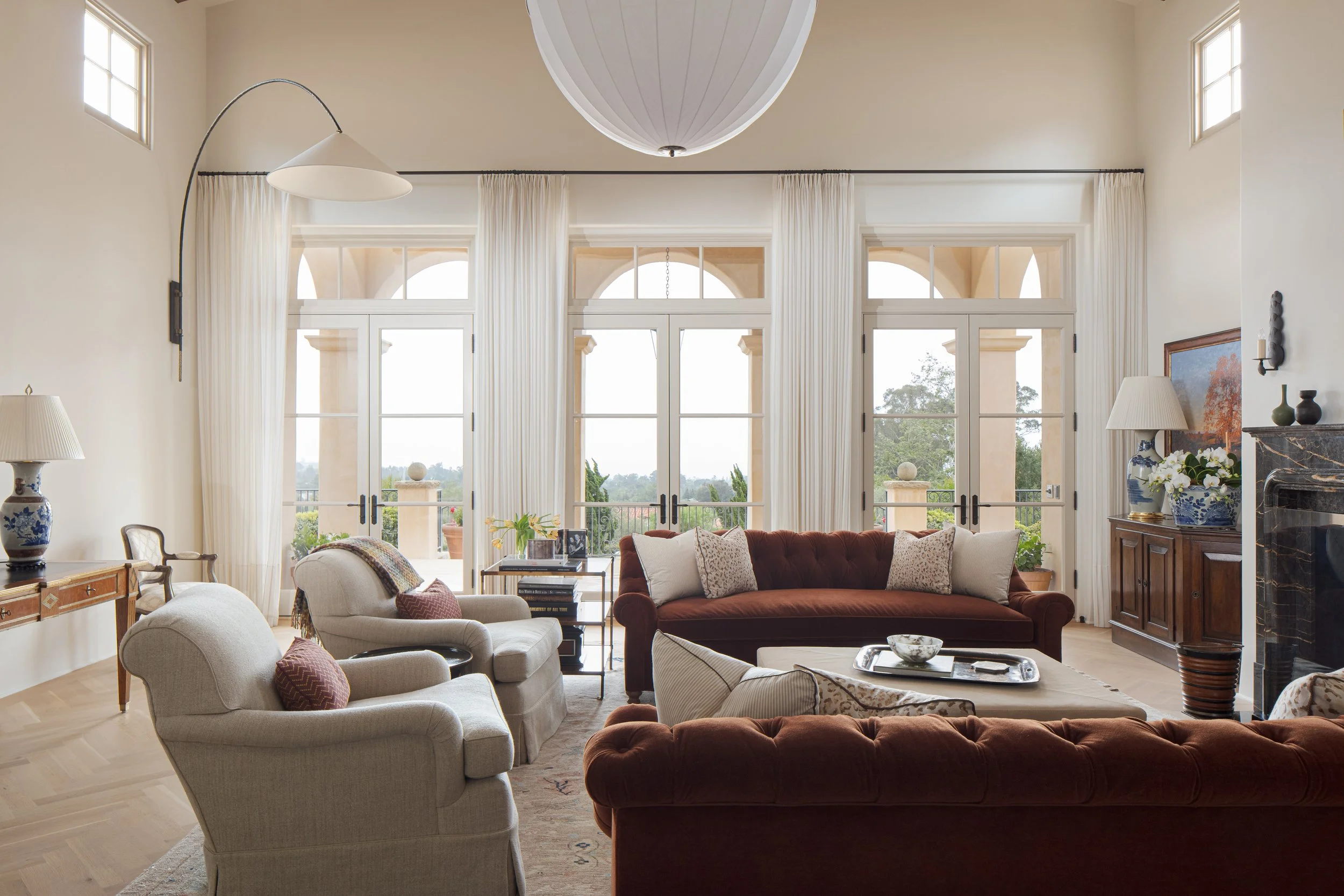 Living room with arched French doors, burgundy tufted sofas, cream armchairs and sheer curtains looking out to the hills