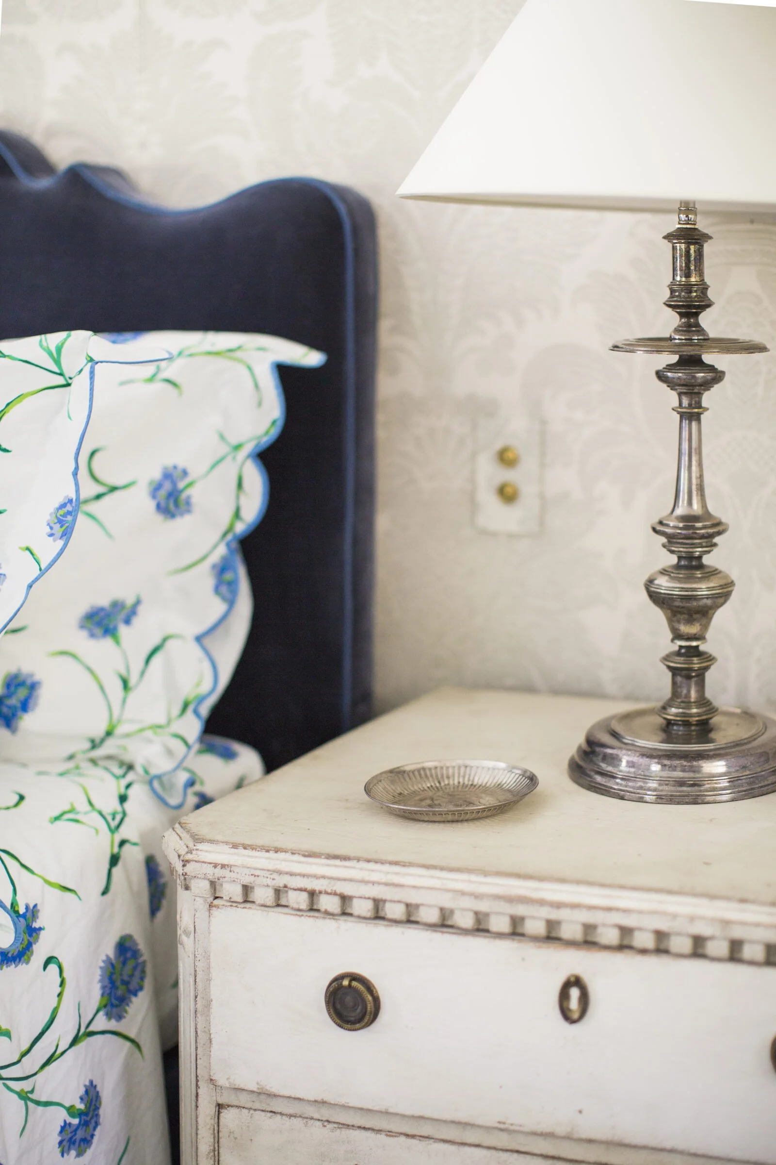 Bedside detail with a navy velvet headboard, floral scalloped bedding, a silver candlestick lamp and a painted Swedish-style nightstand
