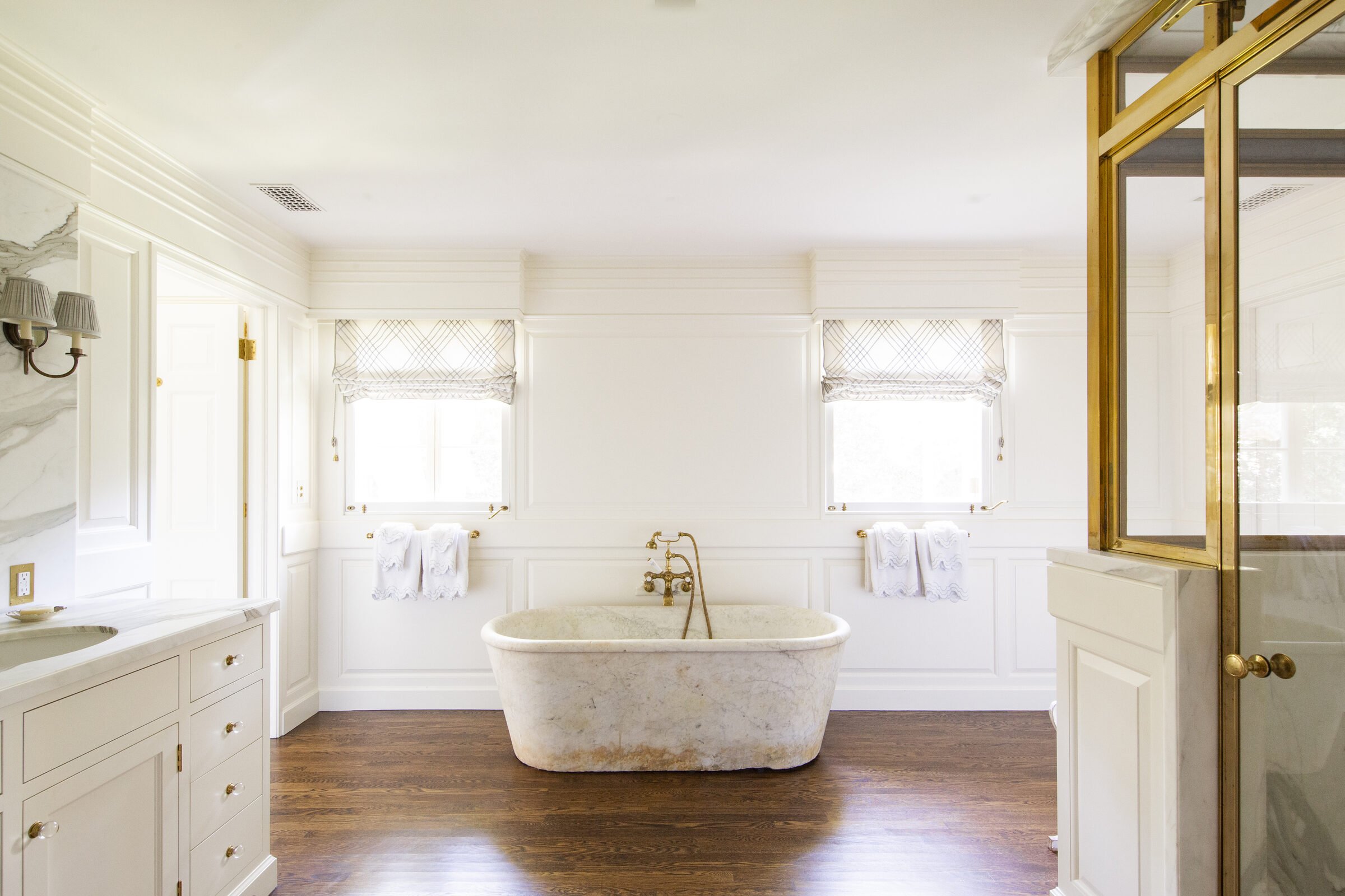 Primary bath with a freestanding stone tub, white paneled walls, a brass-framed marble shower and a wood floor