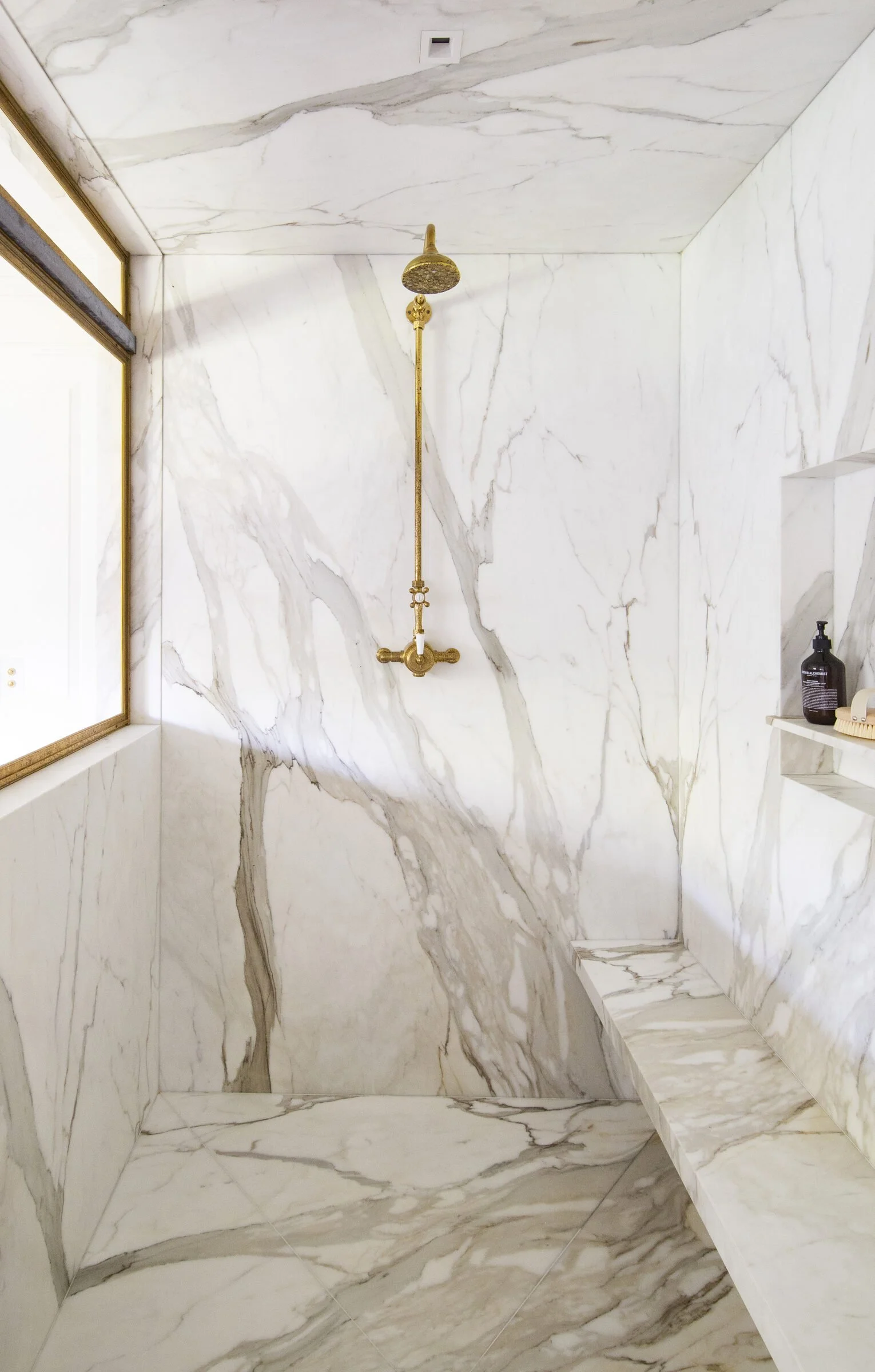 Marble-lined shower with a brass rain shower fixture, a stone bench and a recessed niche