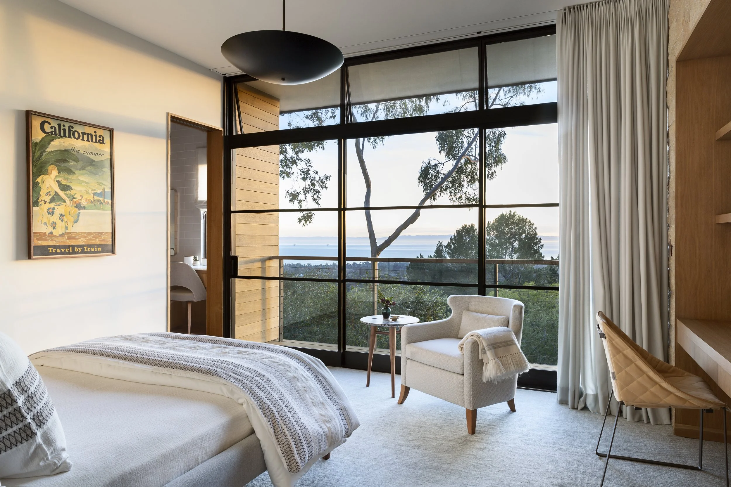 Bedroom with a wall of steel-framed windows overlooking the hills, a white-linen bed, a cream armchair and a vintage California travel poster