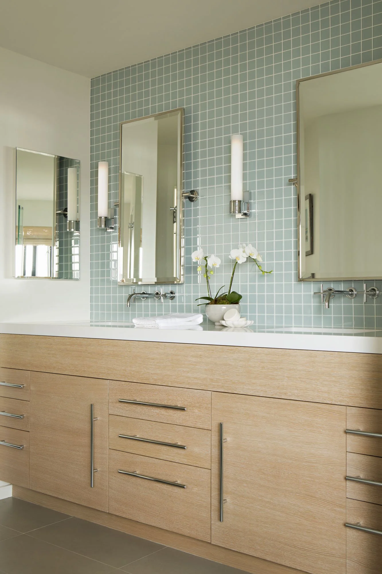 Bathroom with a light oak double vanity, pale blue glass tile, wall-mounted faucets and tall mirrors