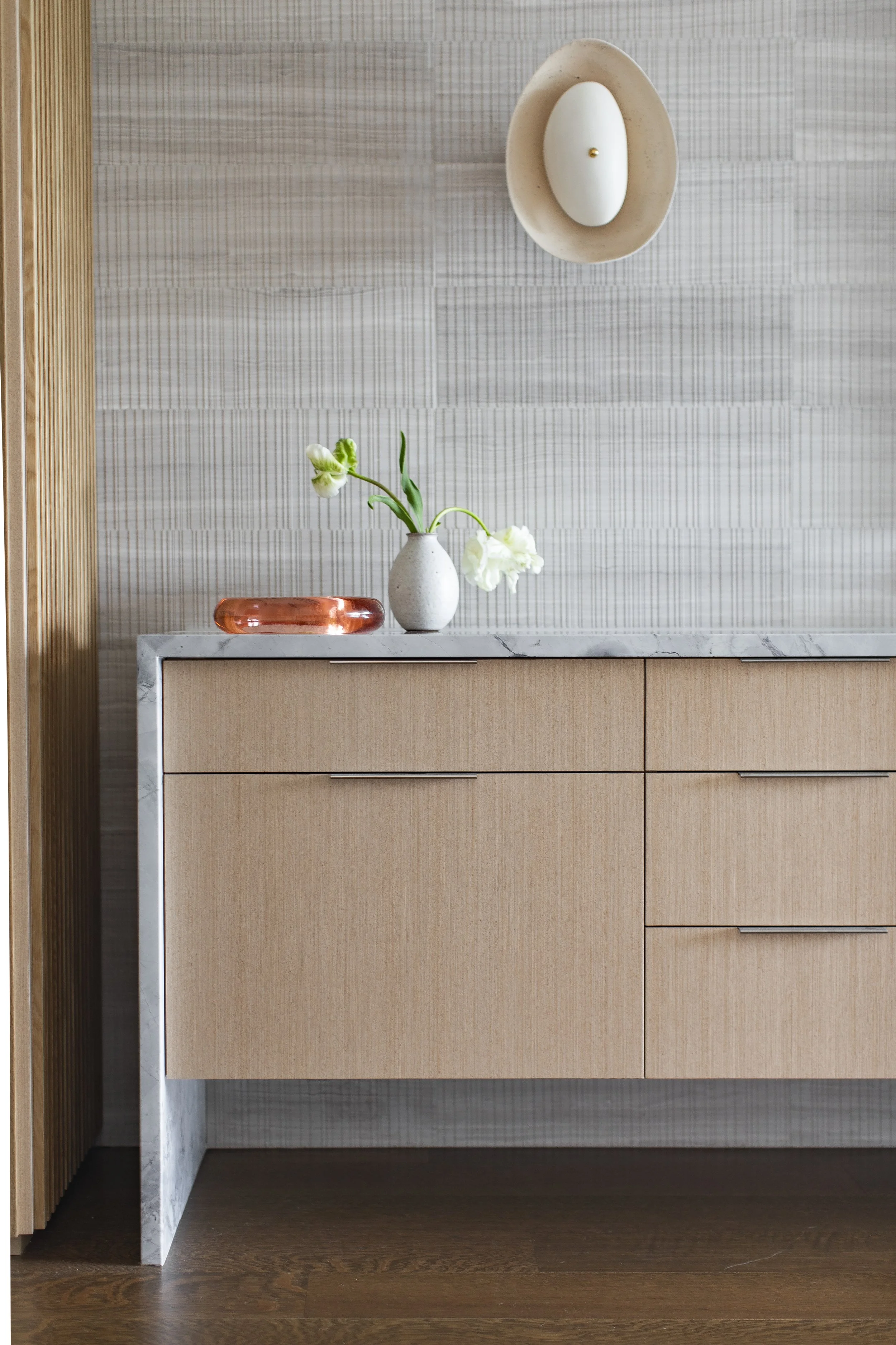 Oak vanity with a stone top and a vase of white flowers against ribbed wallpaper