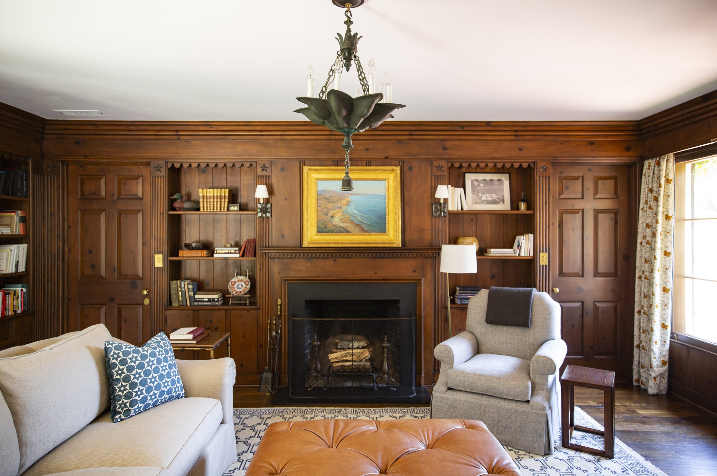 Pine-paneled library with a fireplace, built-in bookshelves, a coastal landscape painting, a gray slipcovered chair and a tufted leather ottoman