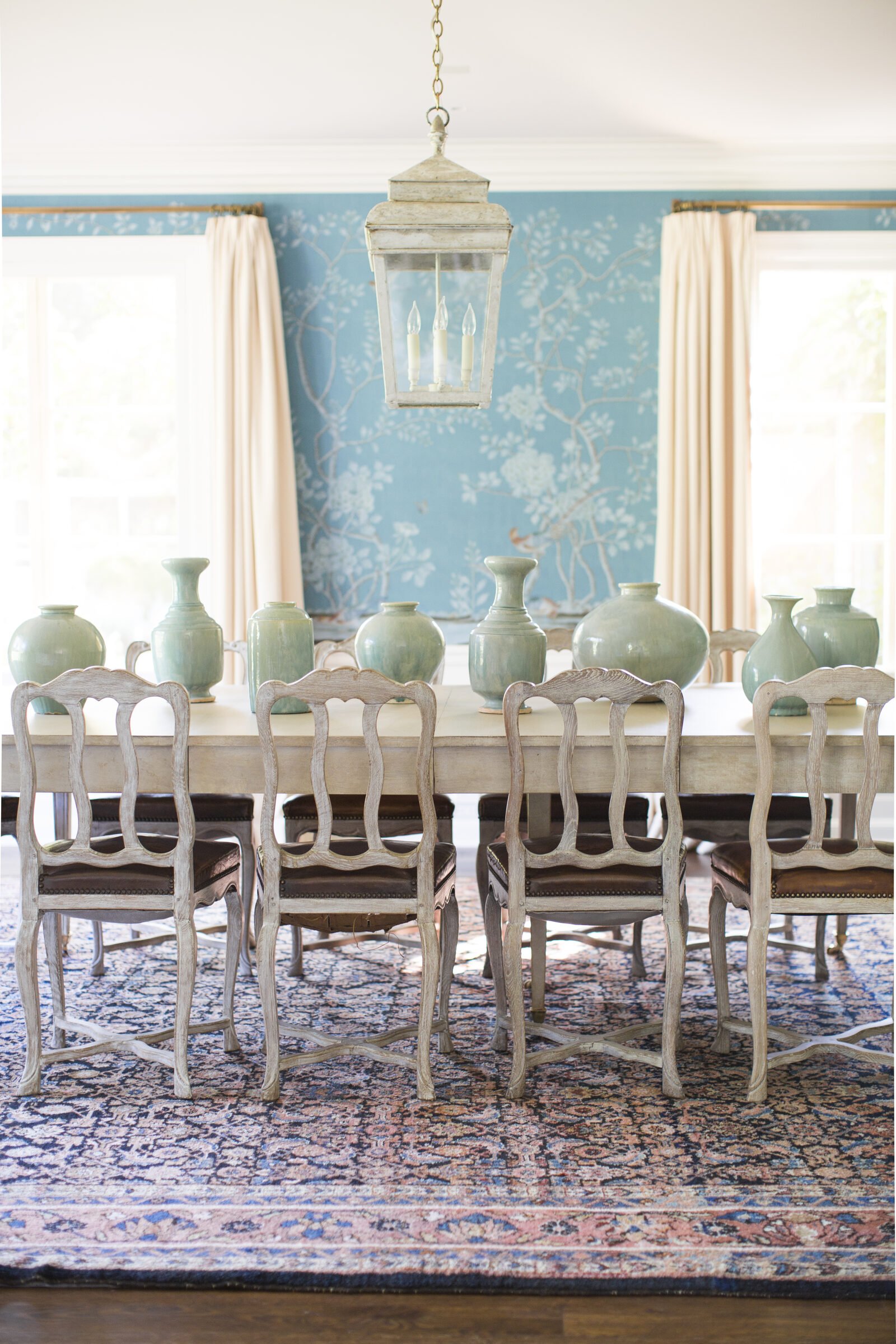 Dining room with blue chinoiserie wallpaper, a table lined with celadon vases, carved painted chairs, a lantern and a blue antique rug