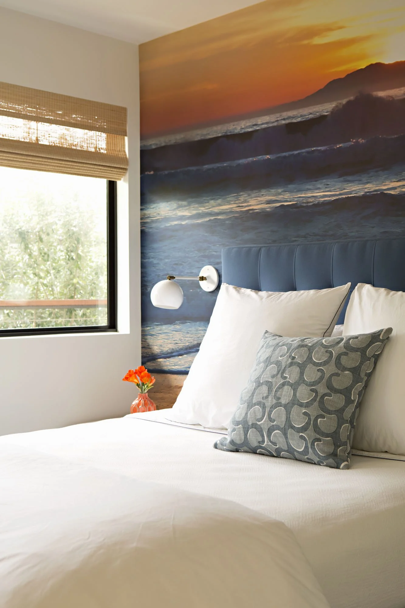 Bed with a blue tufted headboard, a gray patterned pillow and a mural of a sunset sea behind it