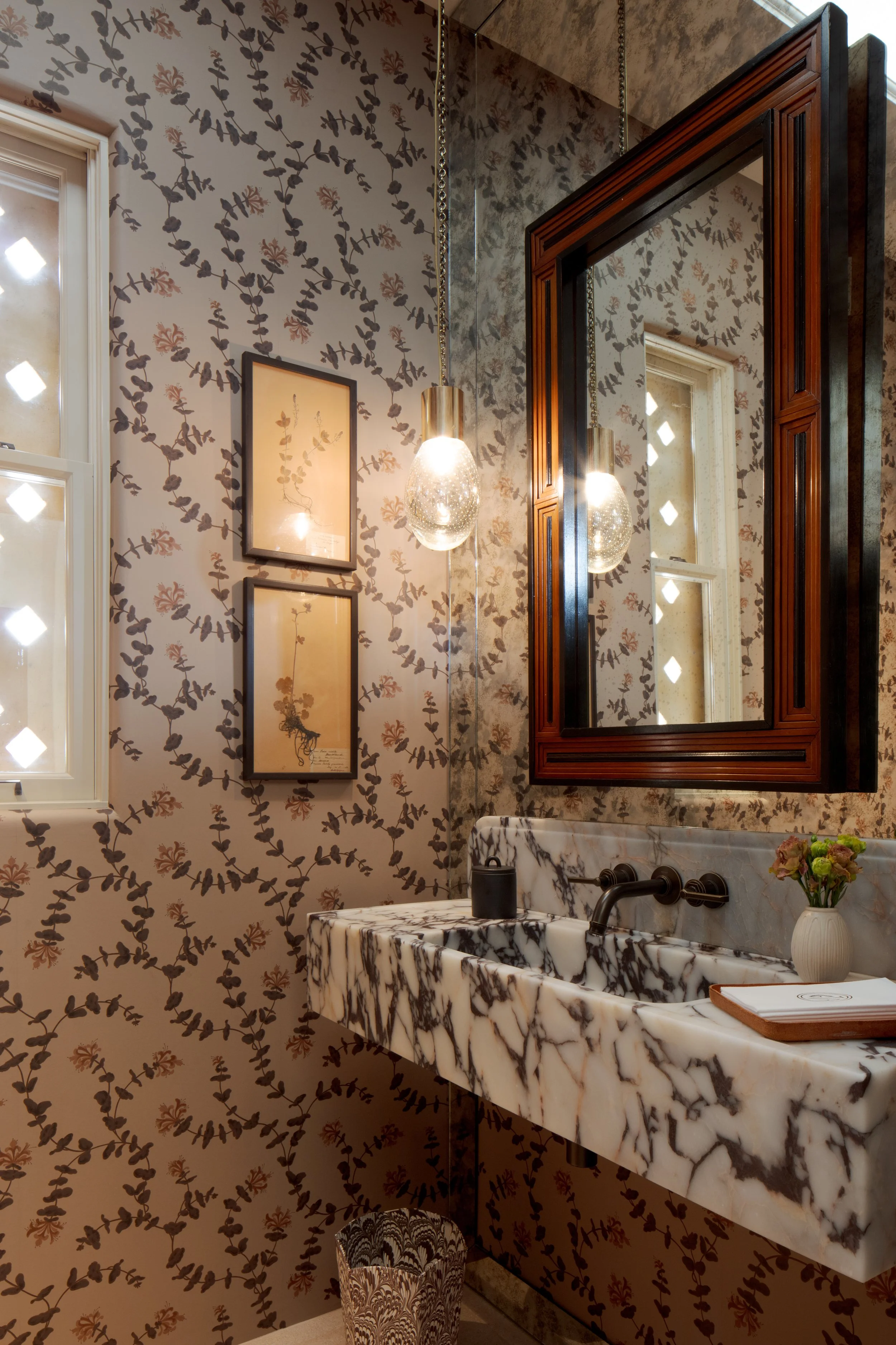 Powder room with floral wallpaper, a marble trough sink, a dark framed mirror and framed botanical prints