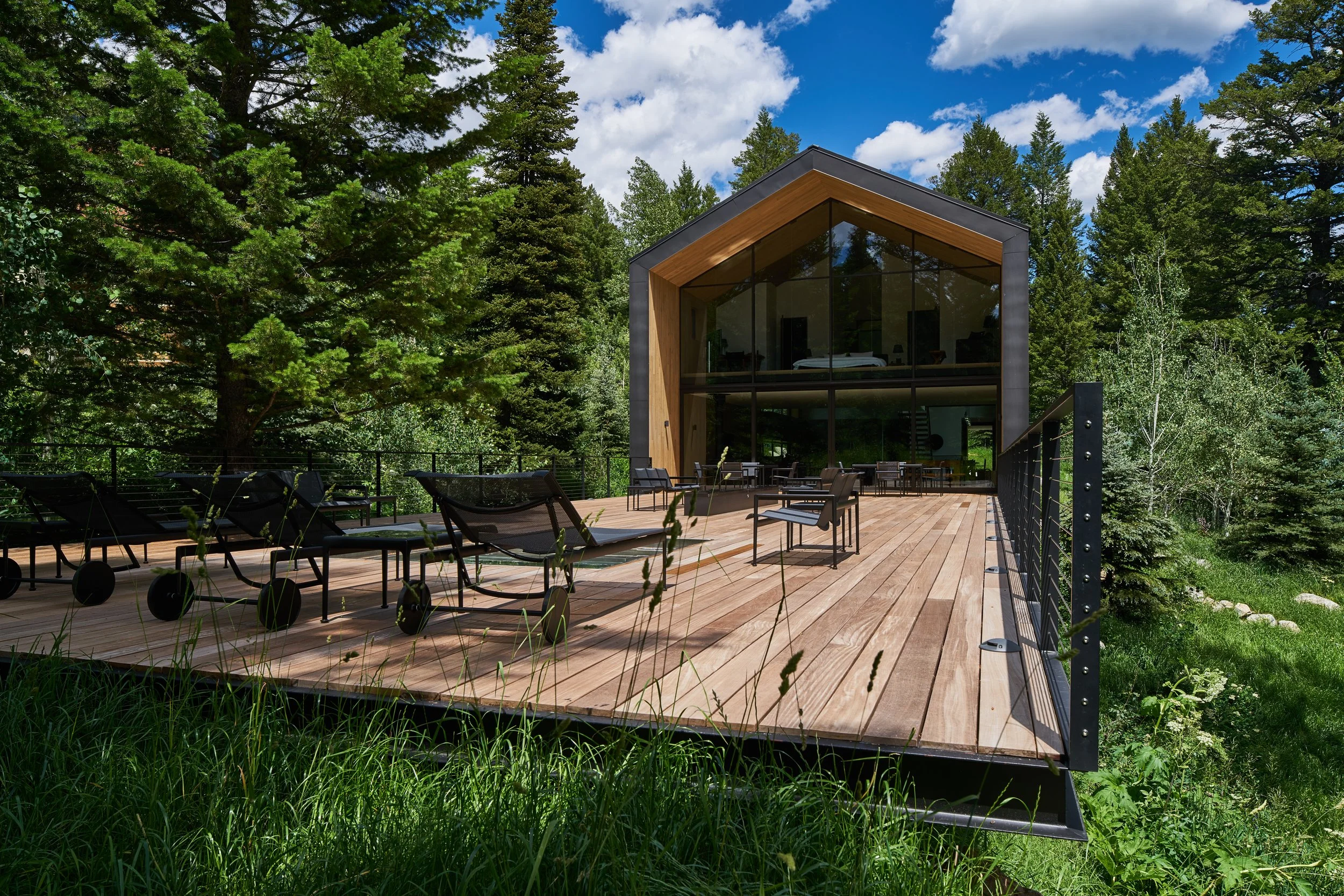 Wood deck with lounge chairs facing the gabled glass house in the trees