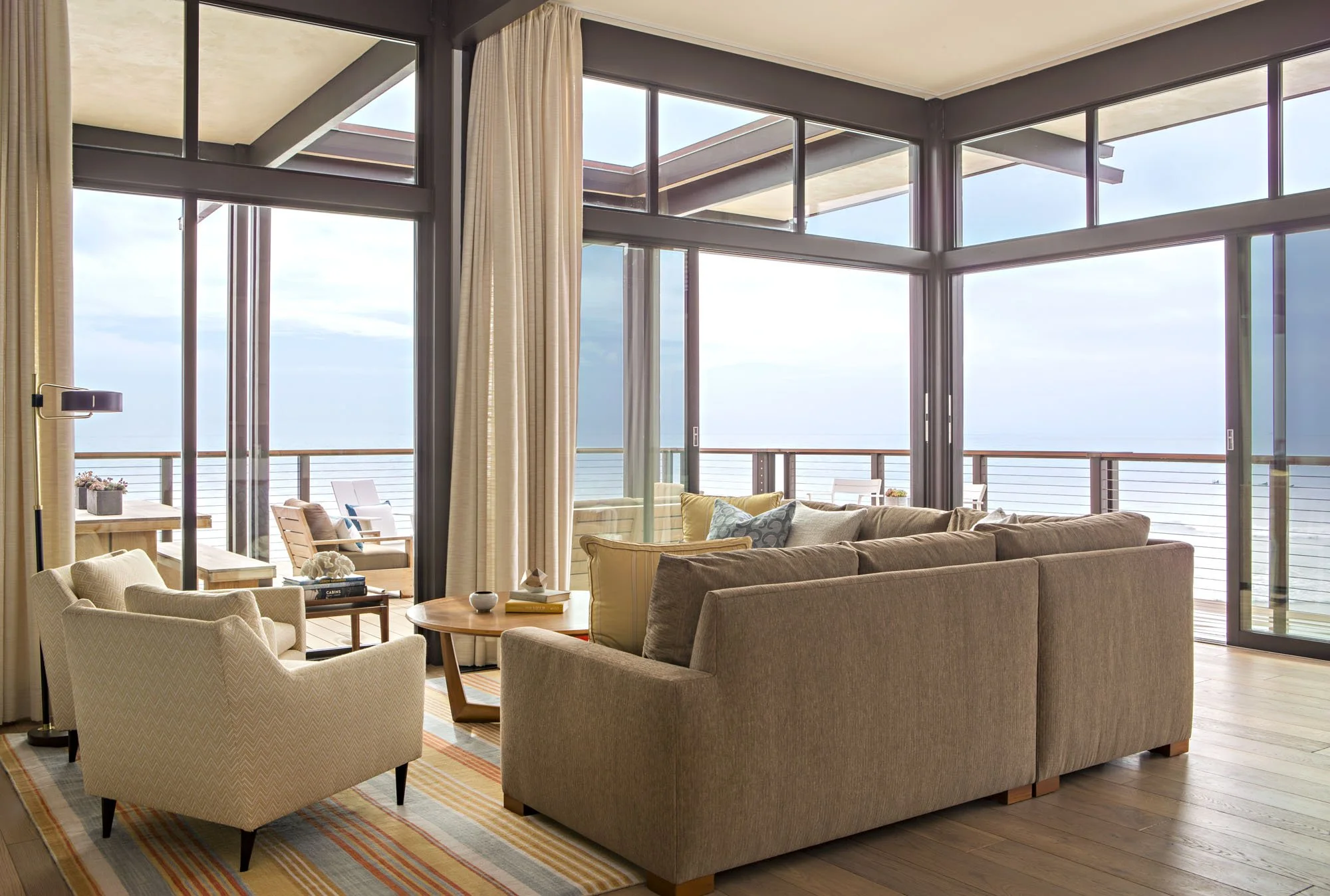 Corner living room with a tan sectional, cream armchairs and walls of glass opening to an ocean-view deck