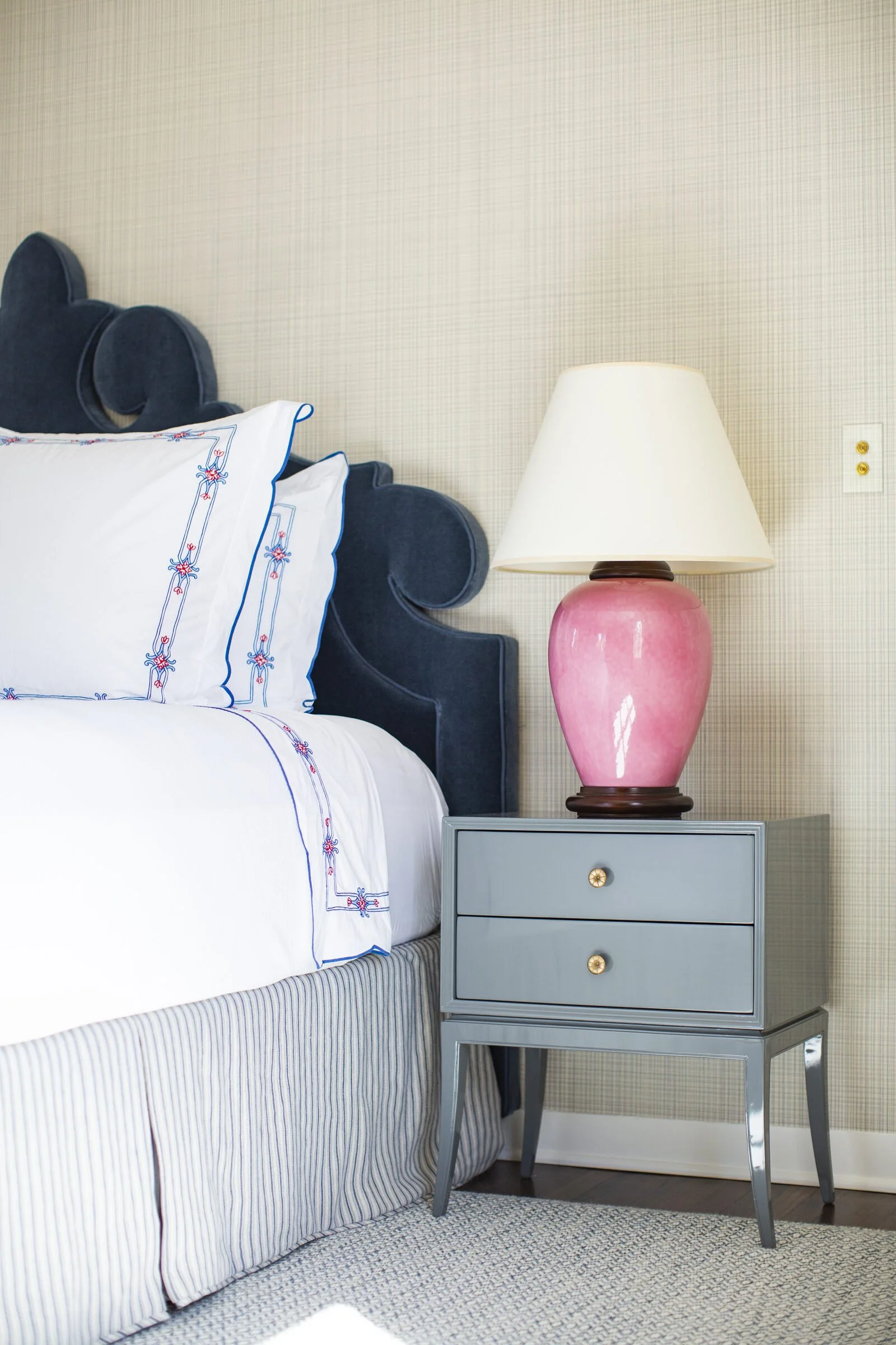 Guest bedroom with a navy velvet headboard, embroidered white linens, a pink ceramic lamp and a blue-gray nightstand