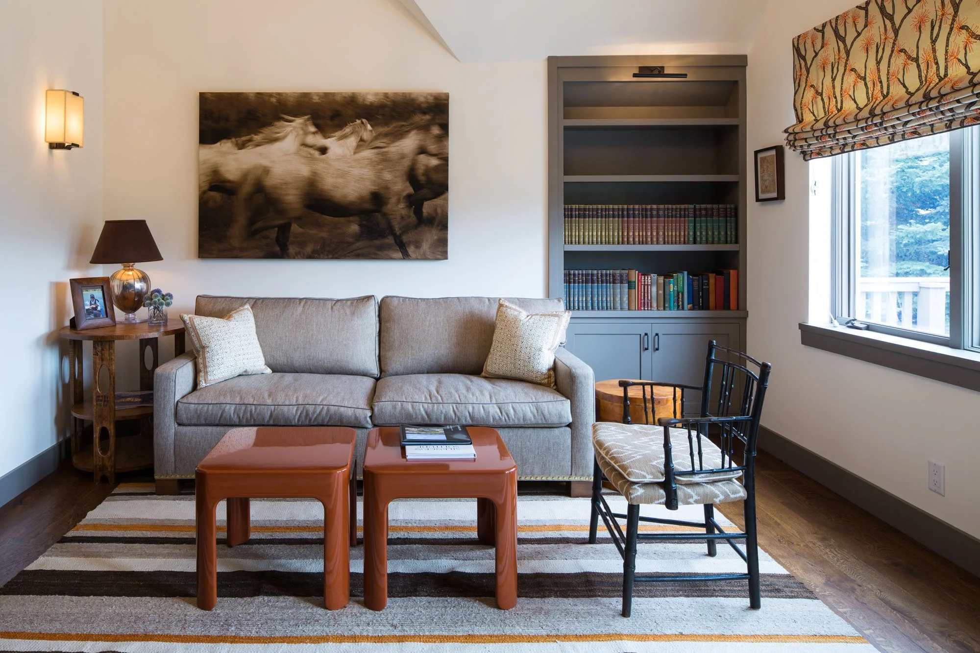 Den with a gray sofa, two rust-colored tables, a framed painting of galloping horses and built-in shelves