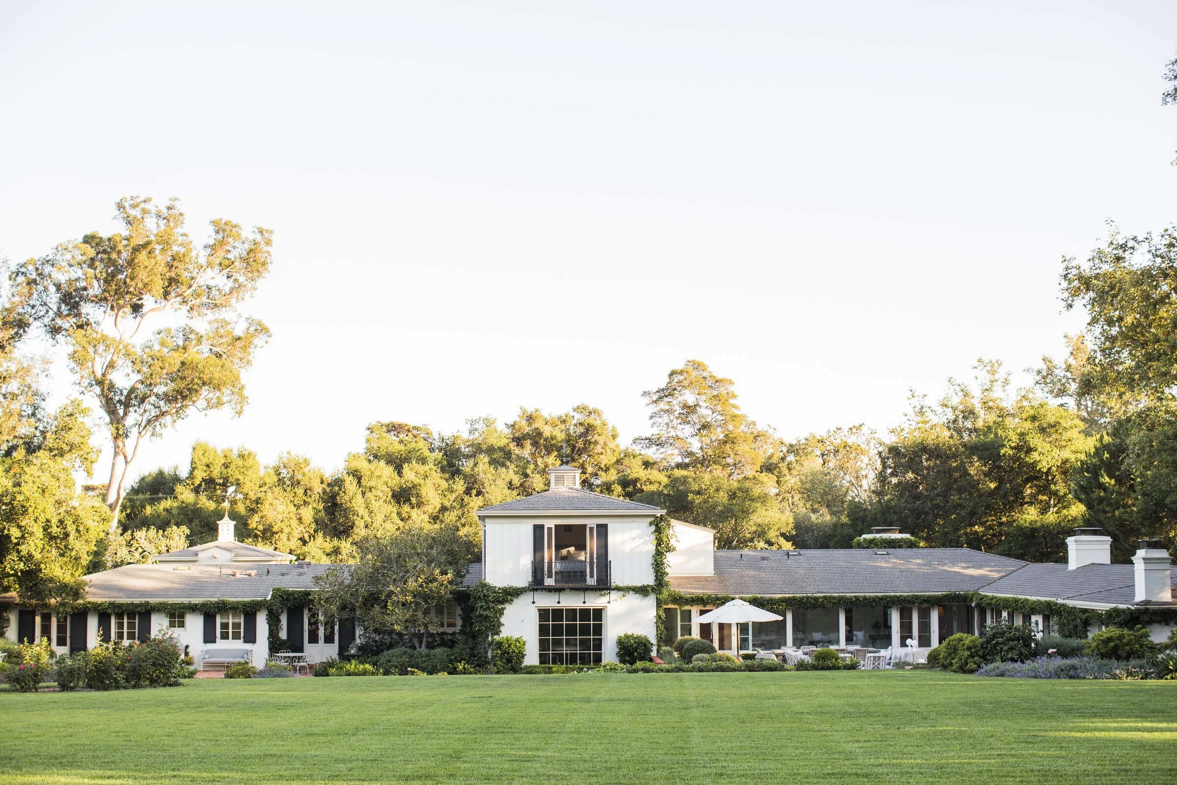 White Montecito house with dark shutters and a gray roof, framed by mature oak trees and a wide green lawn