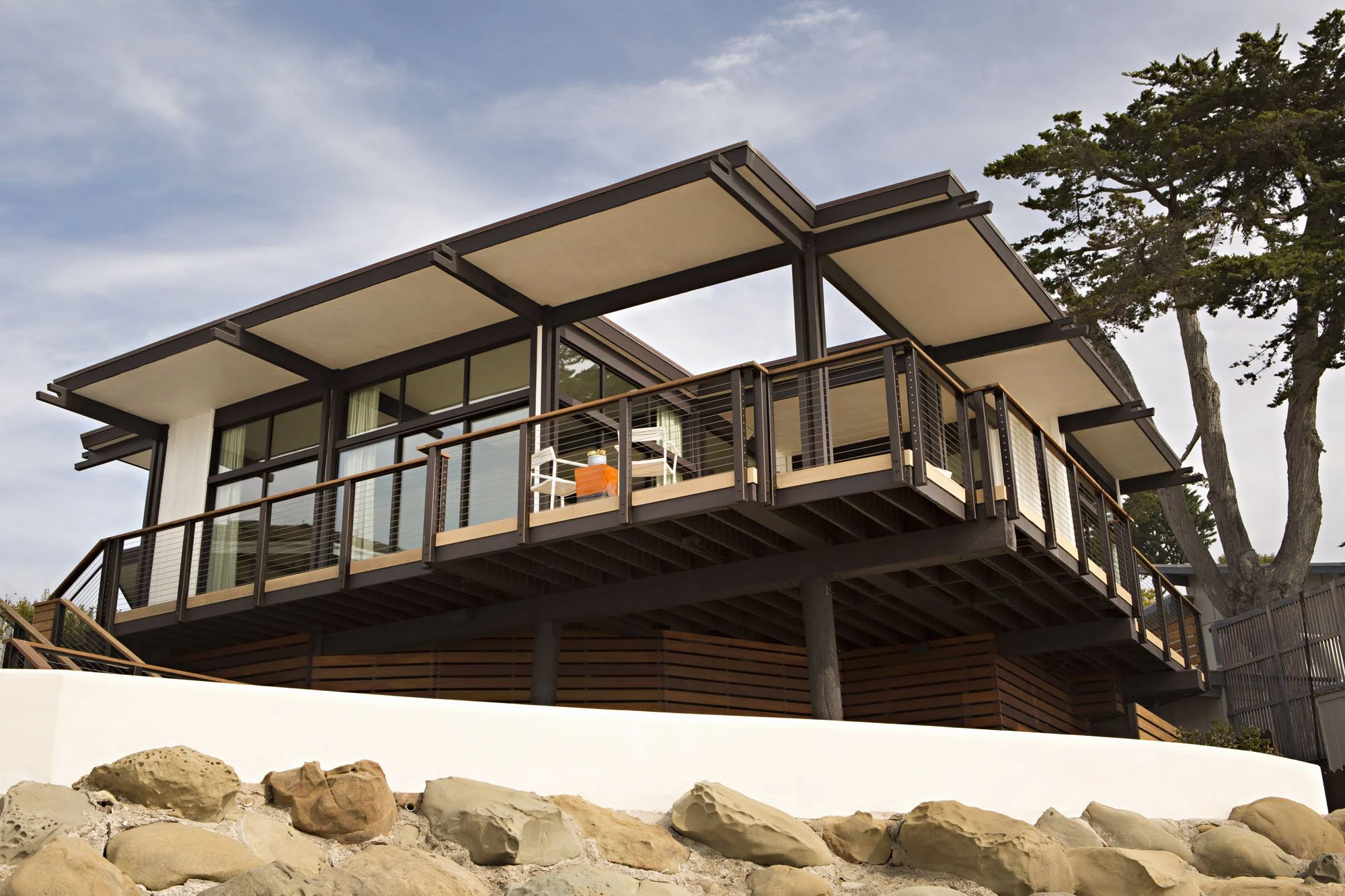Midcentury beach house on stilts with a wide deck, glass walls and a low roof above the rocks at the shoreline