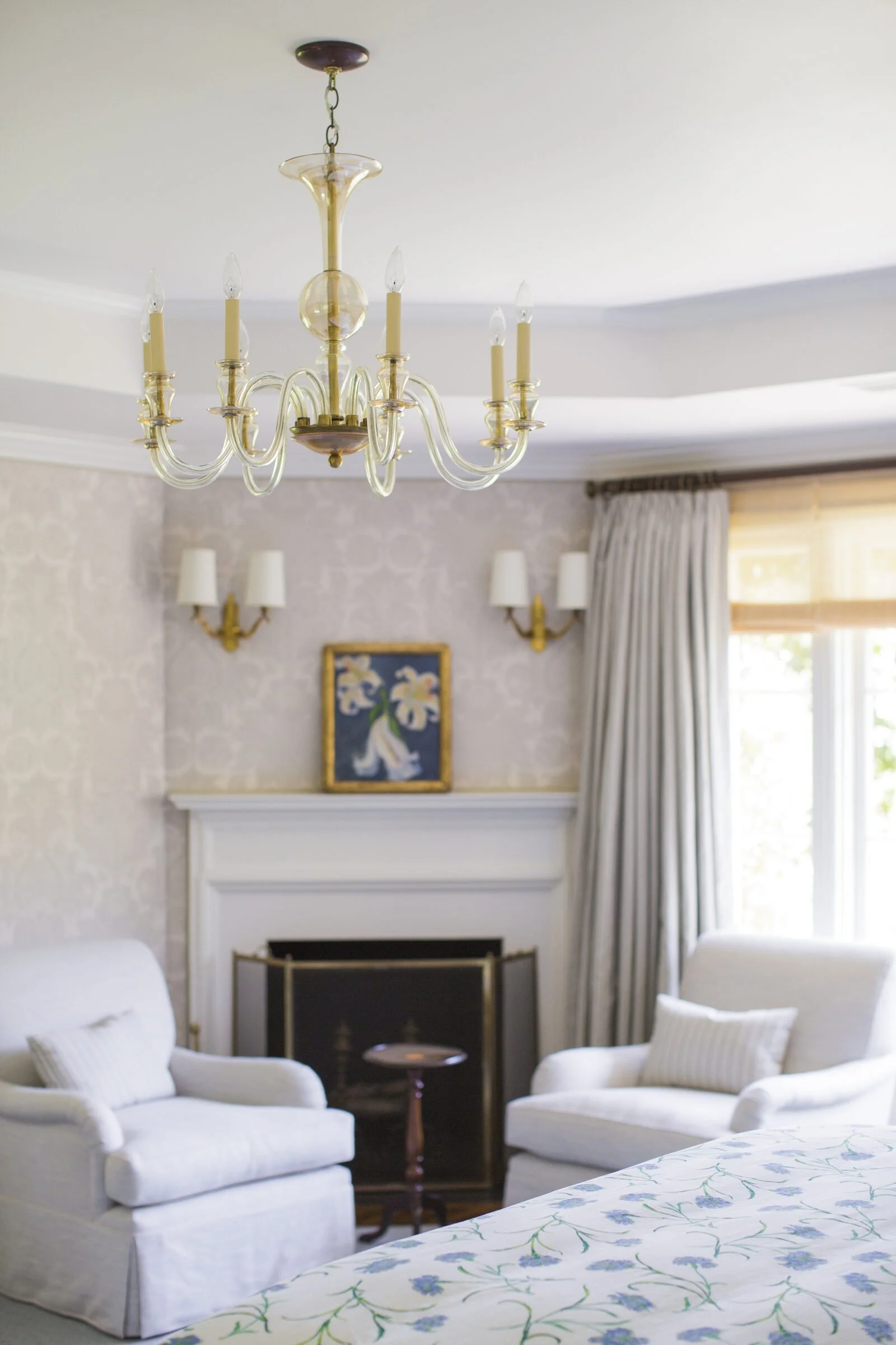 Primary bedroom with a corner fireplace, two white armchairs, a glass chandelier, floral bedding and long gray curtains