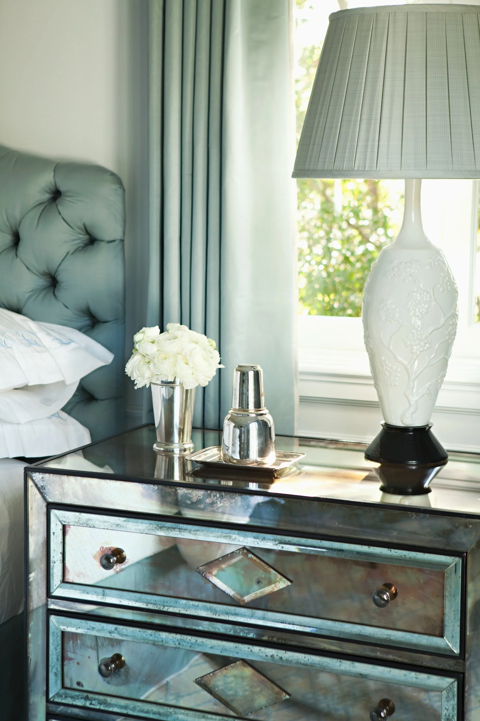 Bedside table with white hydrangeas, a carafe and a carved white lamp beside a teal tufted headboard