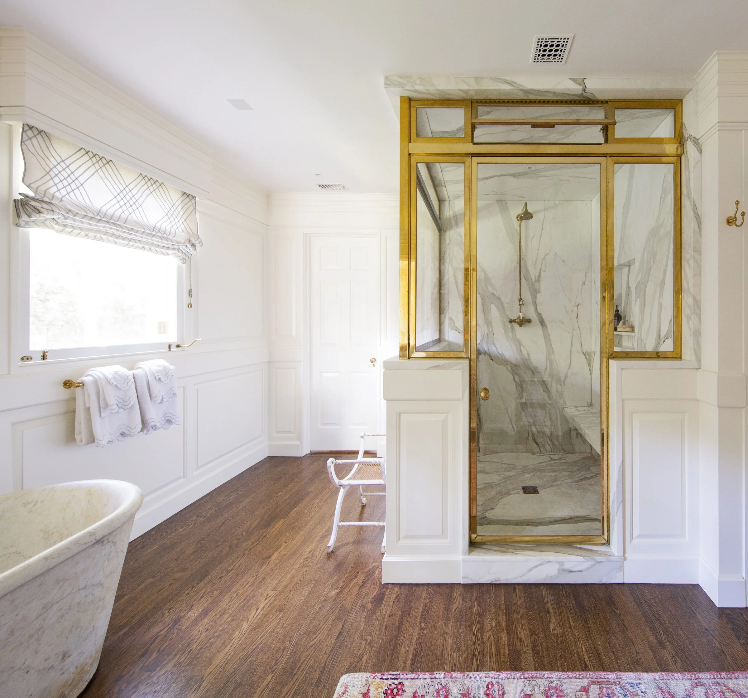 Primary bath with a brass-framed glass and marble shower, white paneling, a plaid Roman shade and a stone tub