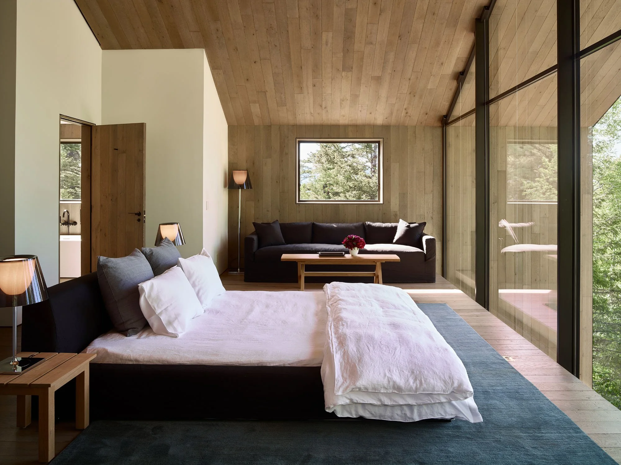 Bedroom with a wood ceiling, dark upholstered bed and a glass wall to the trees