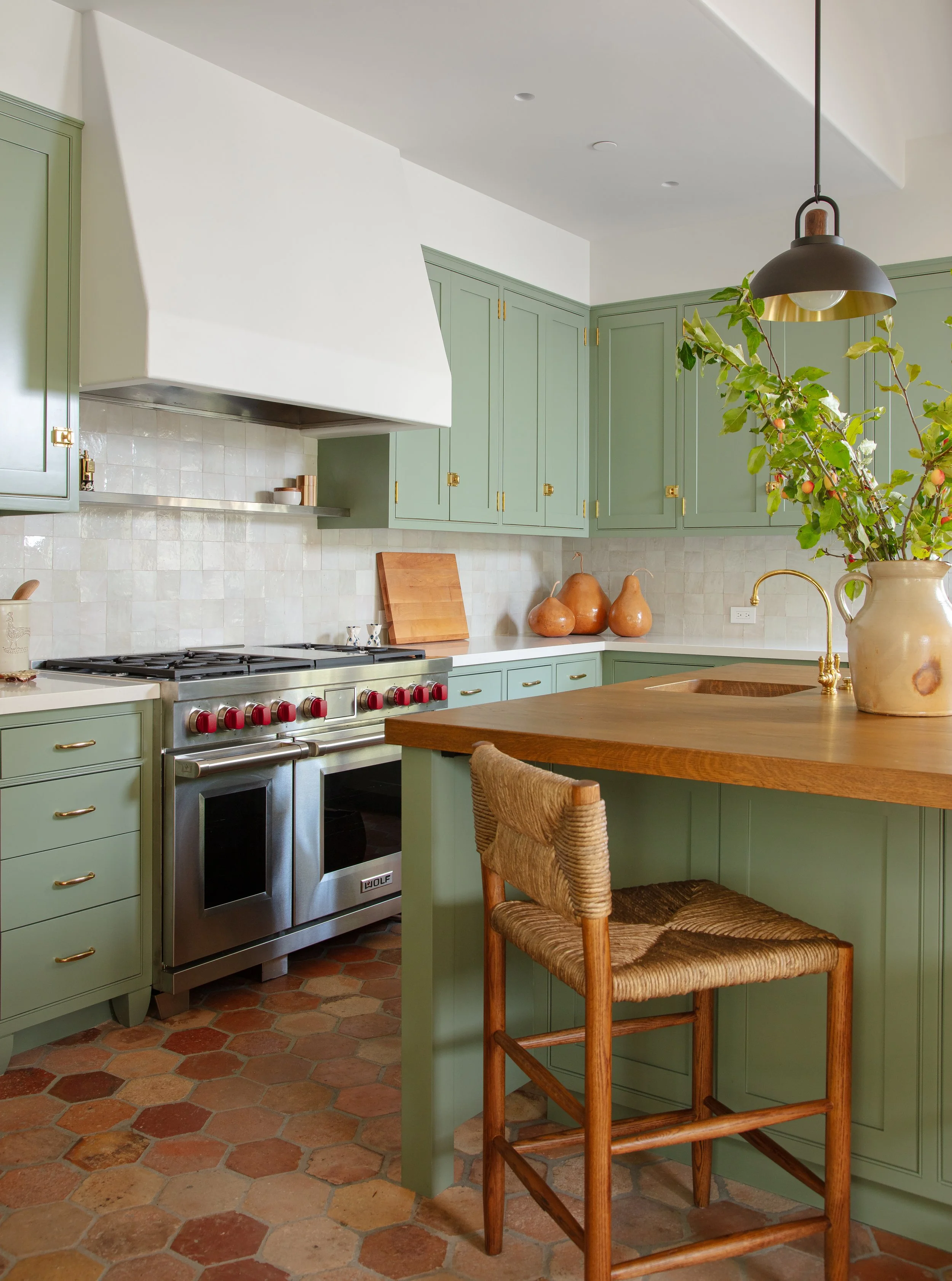 Green kitchen with a stainless steel range, a white plaster hood, a brass faucet and a vase of branches