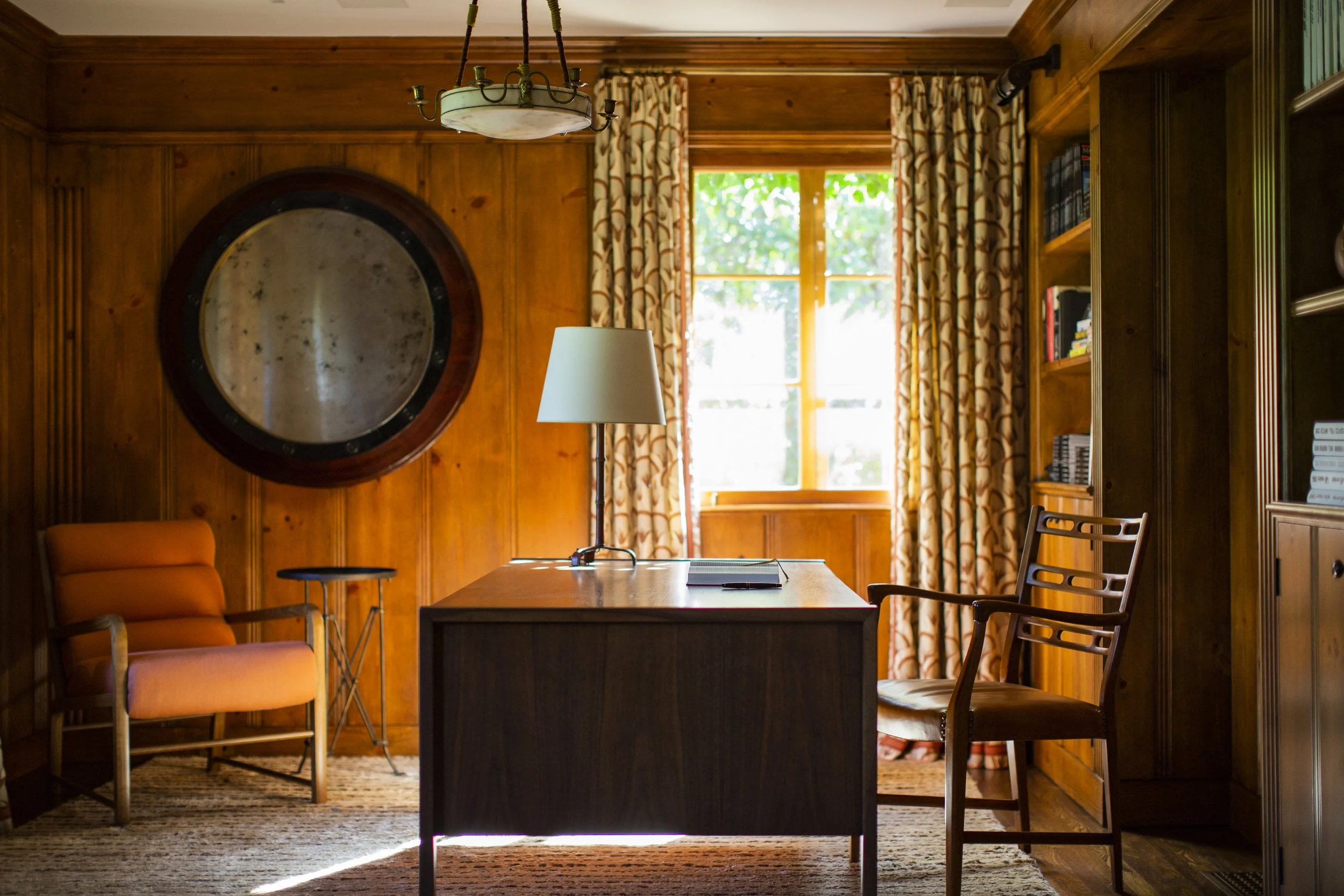 Warm wood-paneled study with a round convex mirror, a dark wood desk, an orange armchair and floral curtains