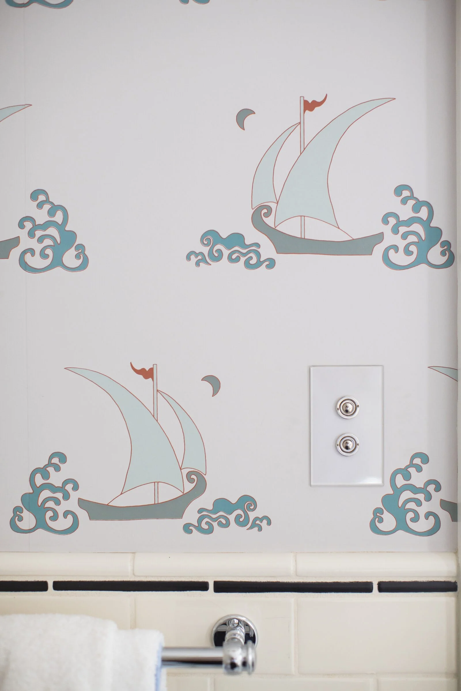 Sailboat and wave wallpaper in soft blue and coral above cream tile with a chrome towel bar