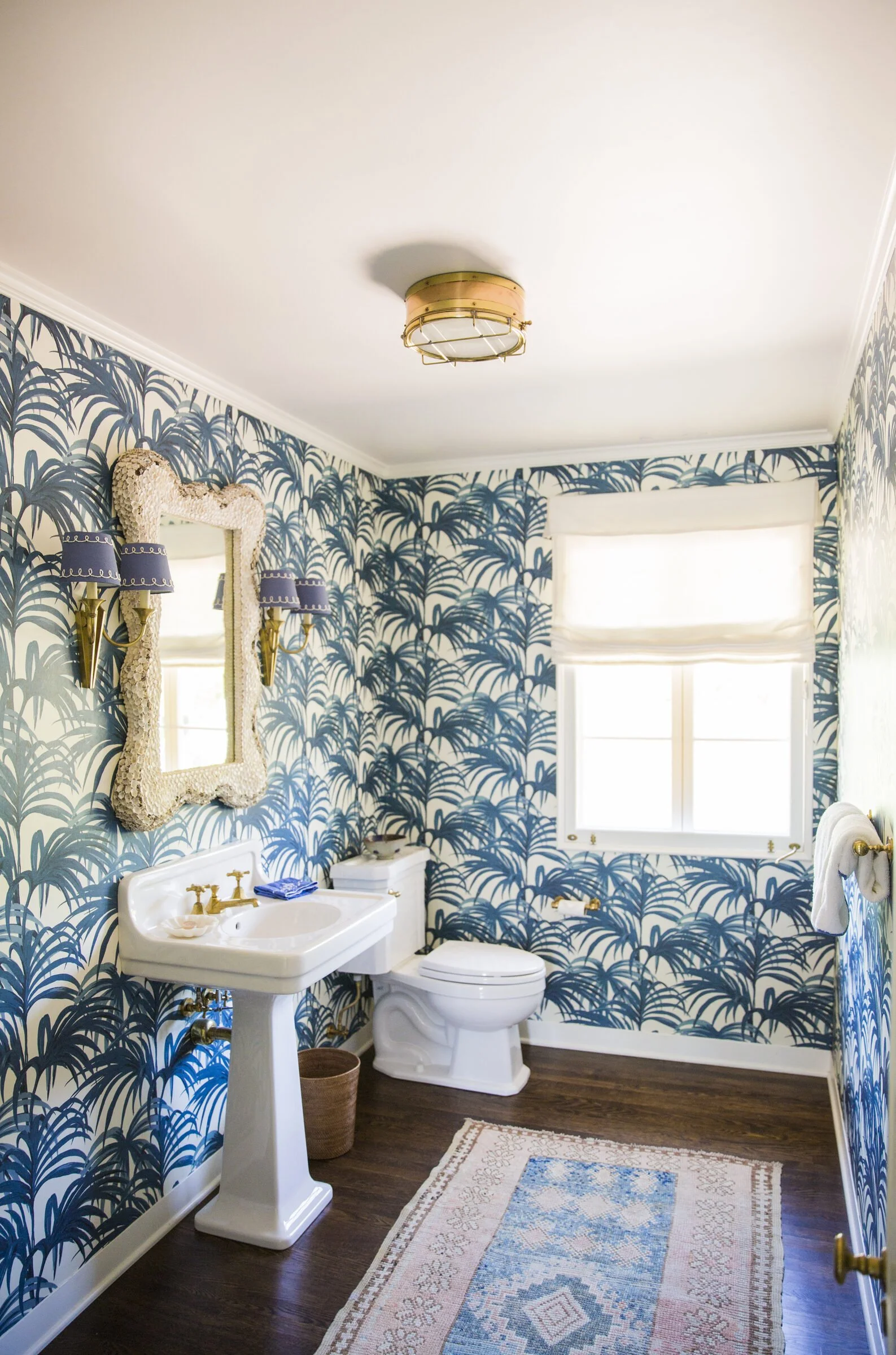 Powder room with blue palm-frond wallpaper, a pedestal sink, a shell-encrusted mirror, brass sconces and a pink and blue runner
