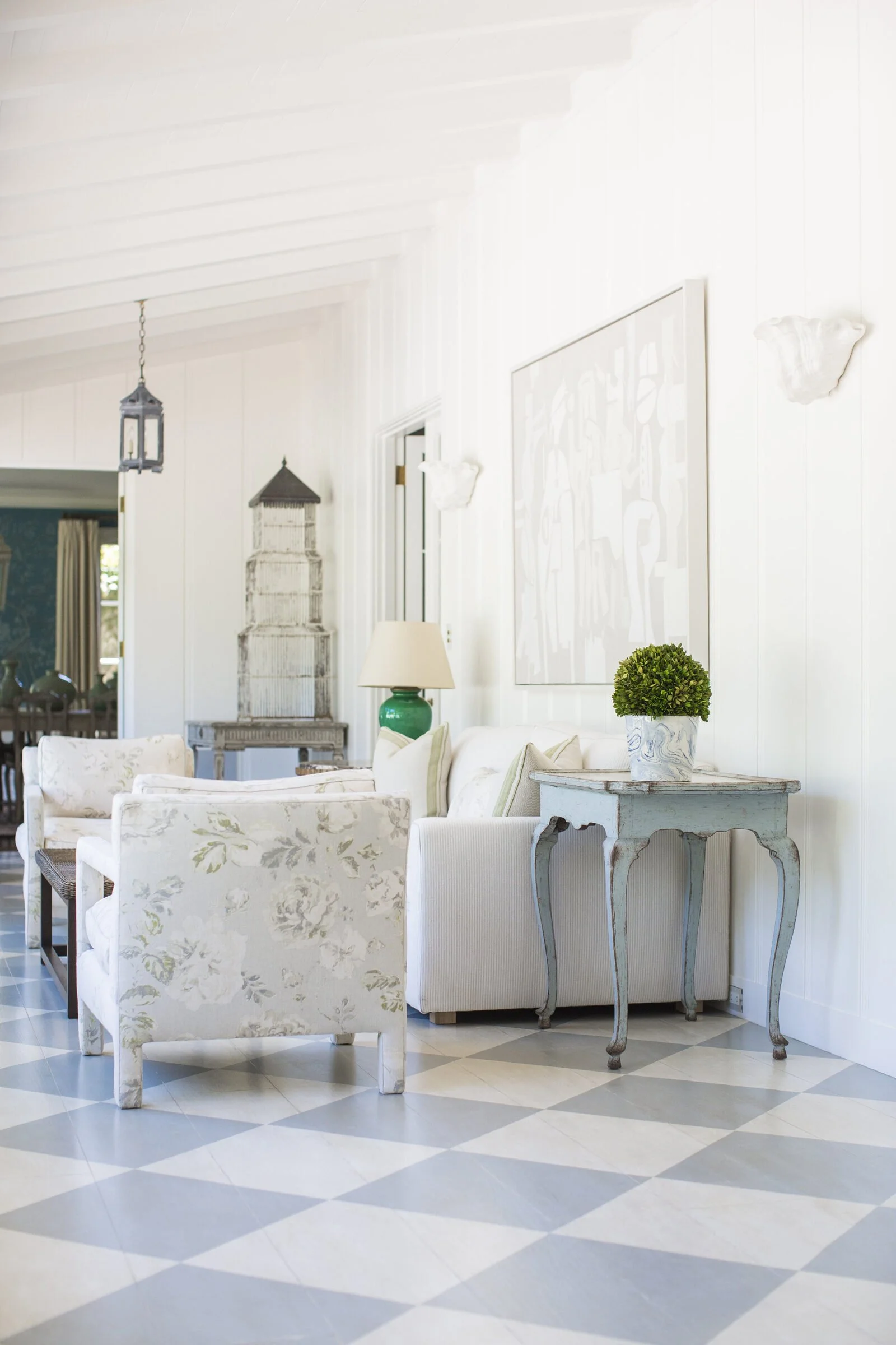 Living room with white paneled walls, a blue-gray and cream painted checkerboard floor, floral armchairs and a pale blue console table