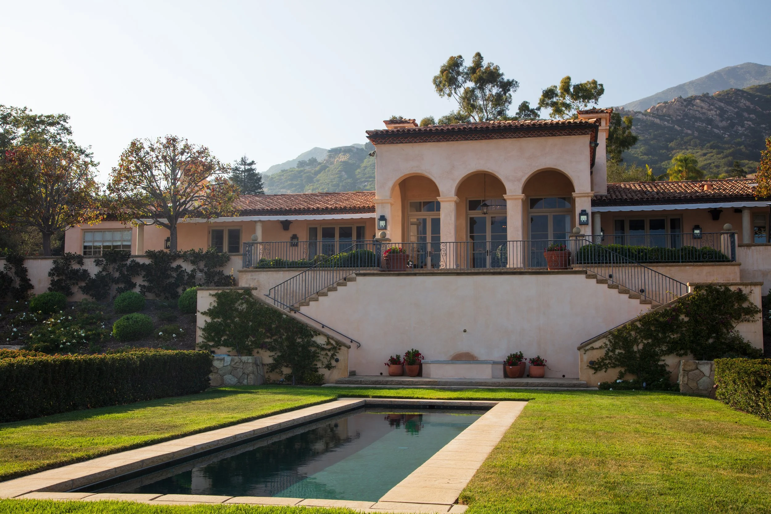 Mediterranean villa with a terracotta tile roof and an arched loggia above a terraced lawn and rectangular pool with mountain views