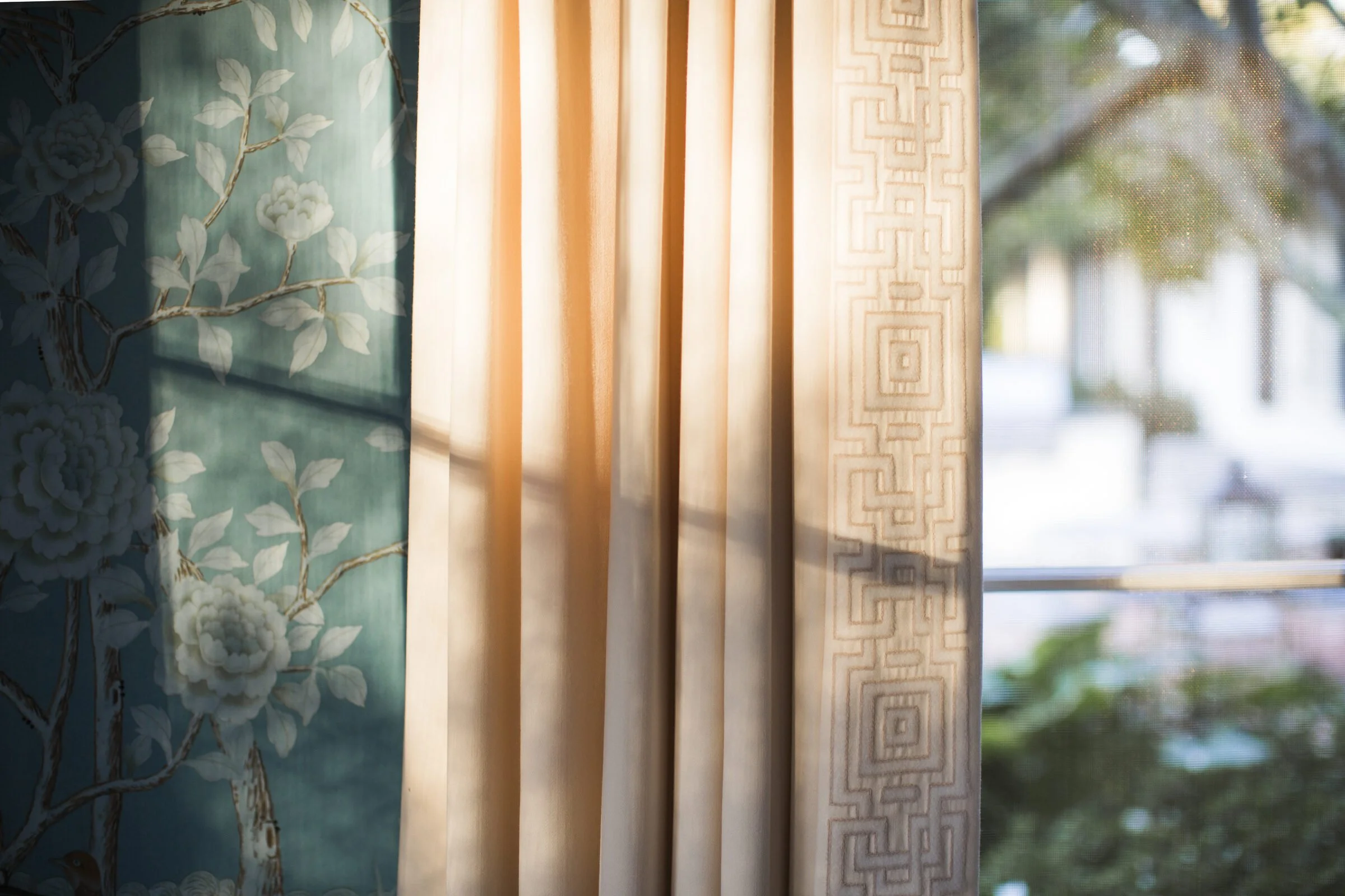 Afternoon light on a cream curtain with a geometric trim beside blue chinoiserie wallpaper