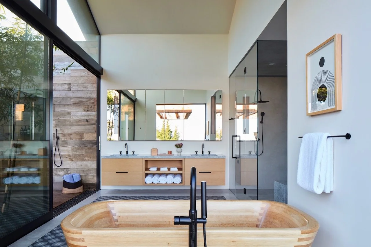 Primary bath with a wooden soaking tub, floating oak double vanity and glass walls opening to a courtyard