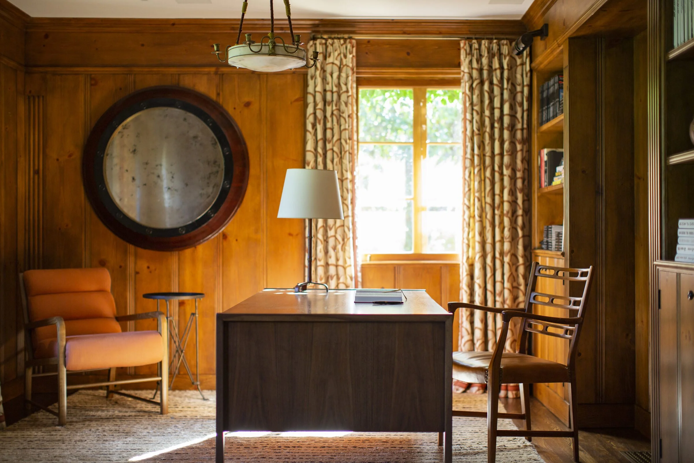 Wood-paneled study with a round antiqued mirror, a dark walnut desk, an orange armchair and printed curtains at a sunny window